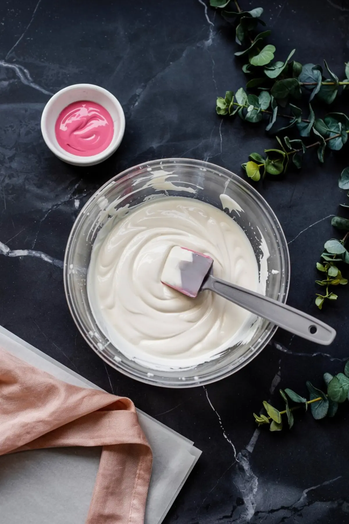 Glass bowl of smooth white royal icing with a spatula, placed beside a small dish of pink icing on a black marble surface with greenery accents.