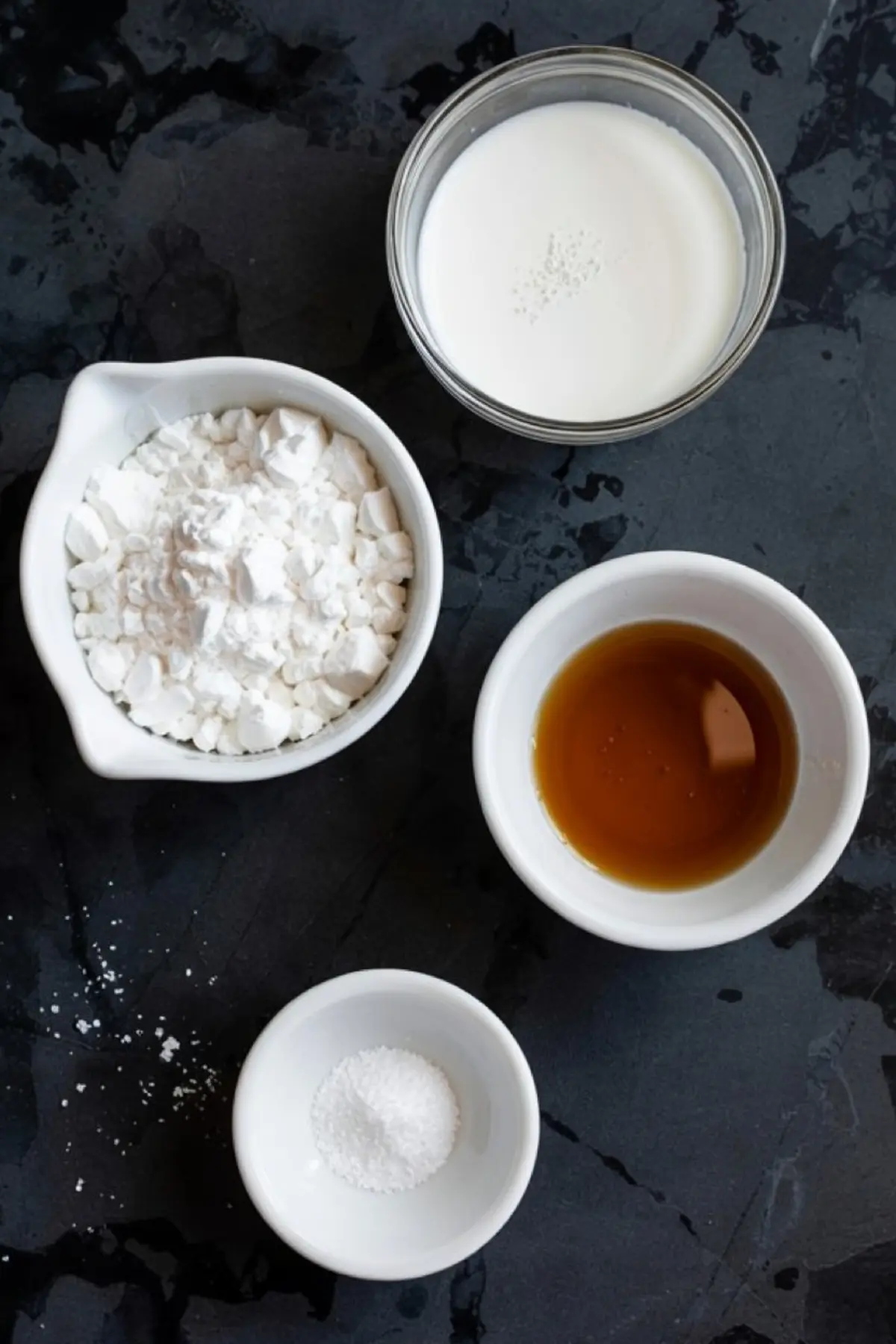 Flat lay of powdered sugar, vanilla extract, milk, and salt in small bowls on a dark textured surface, showing ingredients for powdered sugar frosting.