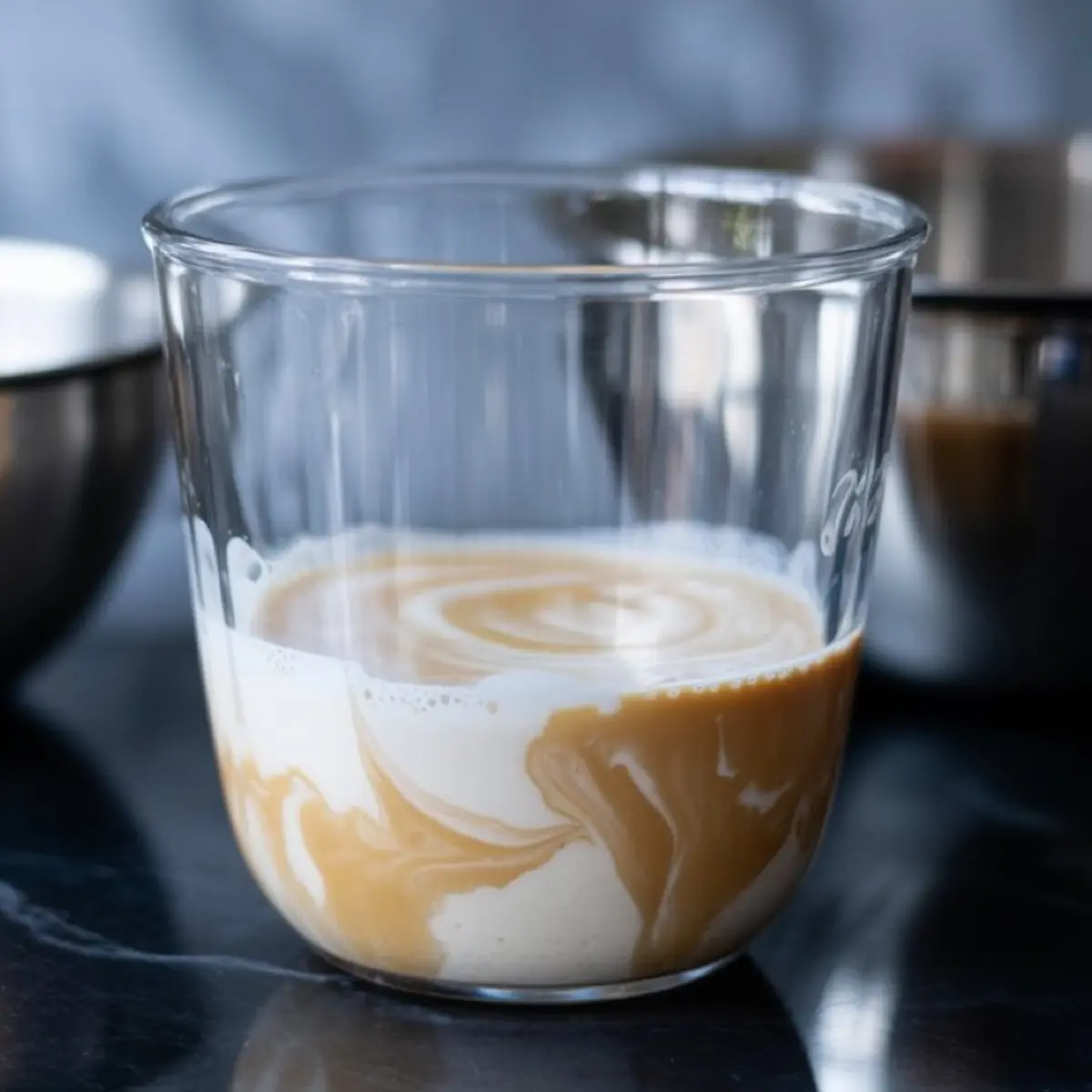 A clear glass with swirling layers of pumpkin purée and cream, showing the blending process for pumpkin cold foam on a dark reflective surface.
