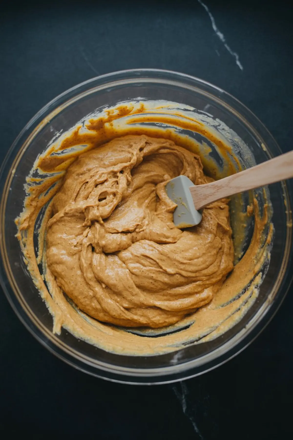Thick pumpkin pancake batter swirled in a glass bowl, mixed with a rubber spatula, showing smooth texture and deep orange color.