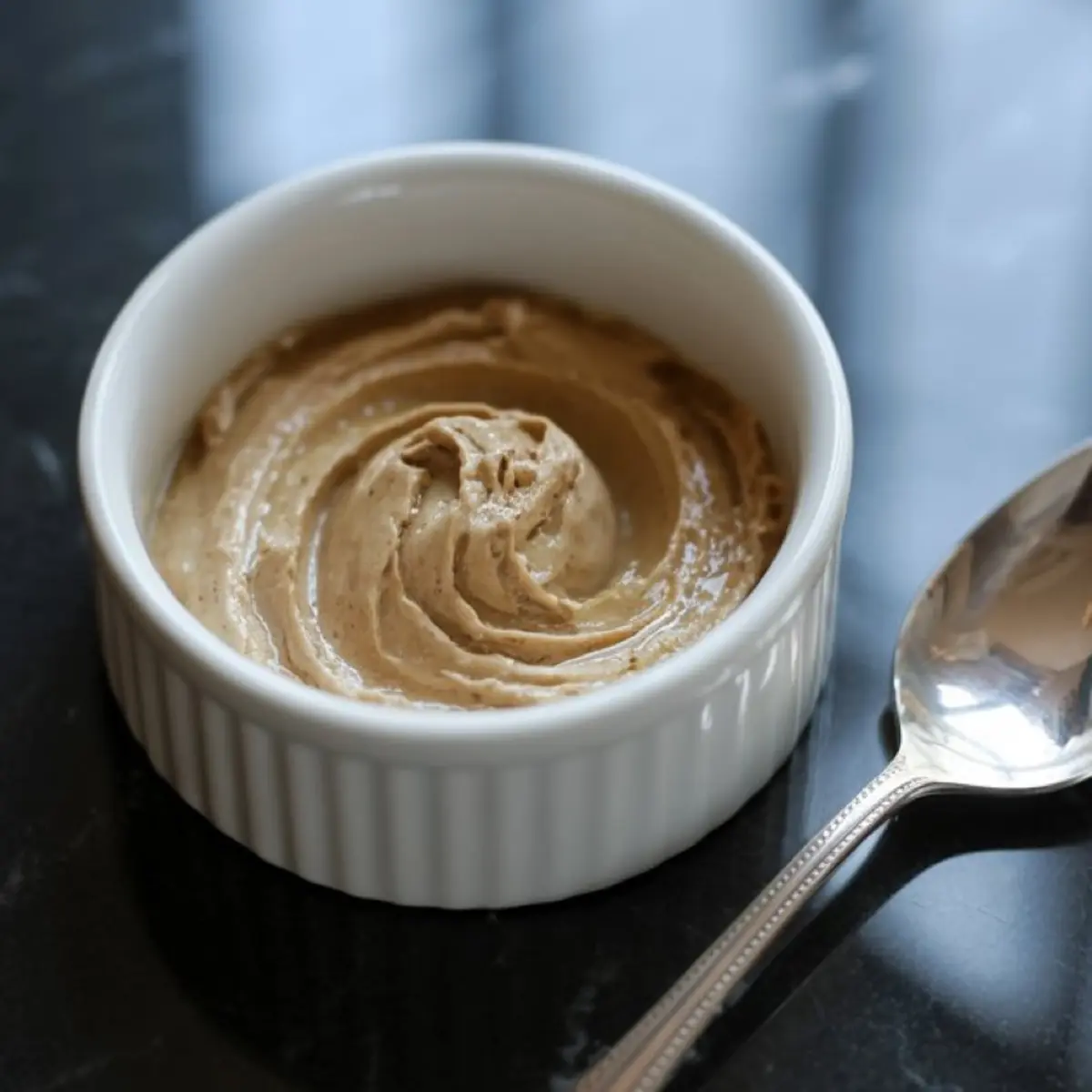 Small white ramekin filled with creamy whipped cinnamon butter, placed next to a silver spoon on a glossy black table.