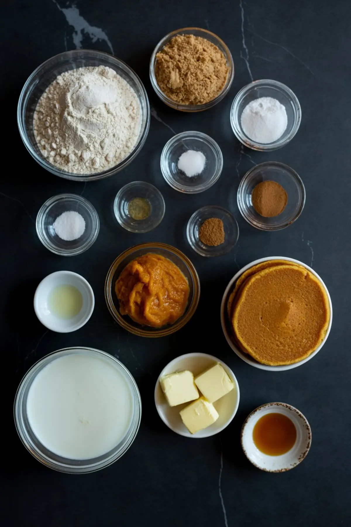 Flat lay of pumpkin pancake ingredients in clear bowls on a dark background, including flour, brown sugar, spices, pumpkin puree, butter, vanilla, and cooked pancakes.