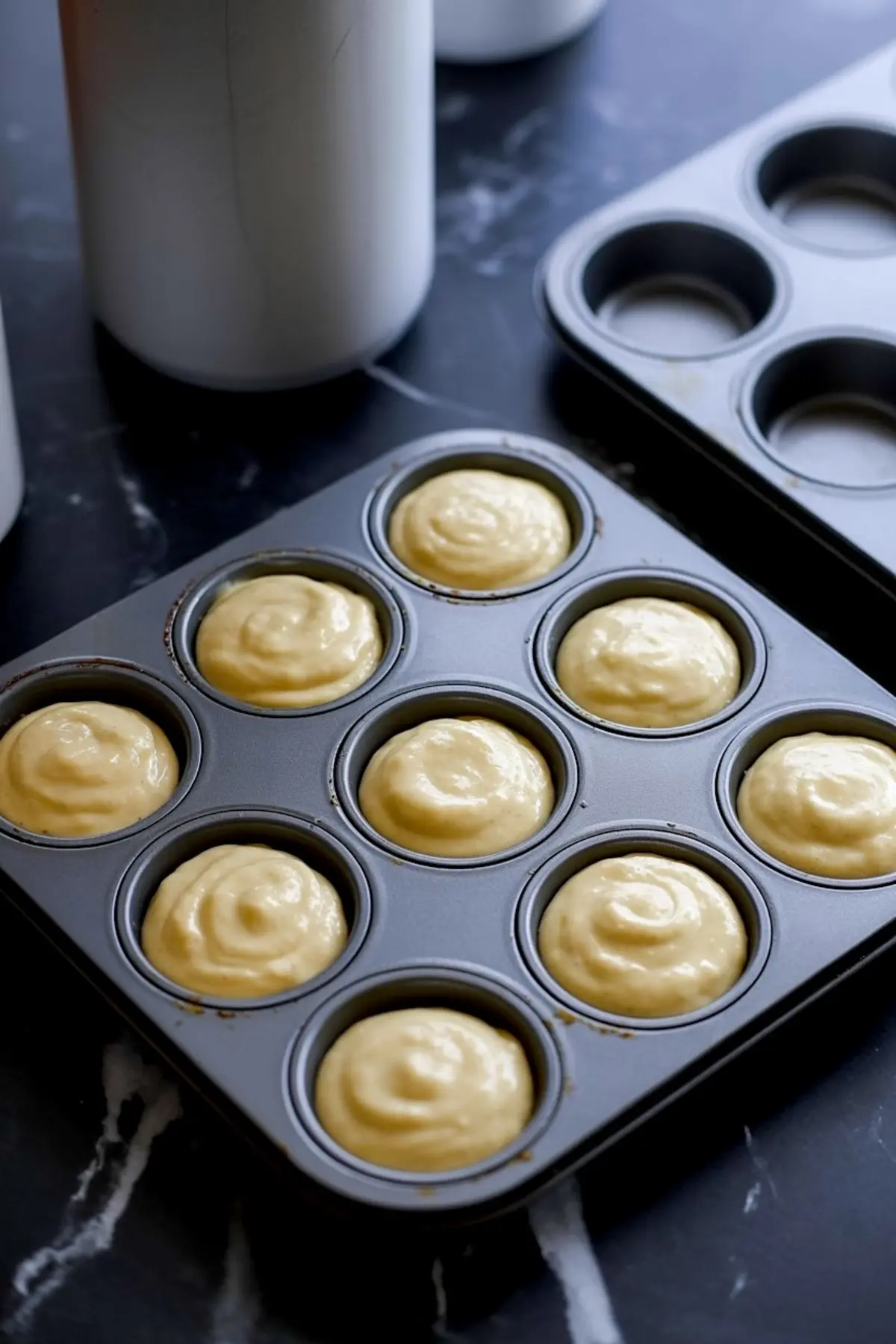 Non-stick muffin tray filled with smooth, light yellow cupcake batter, ready for baking, surrounded by empty molds on a dark countertop.
