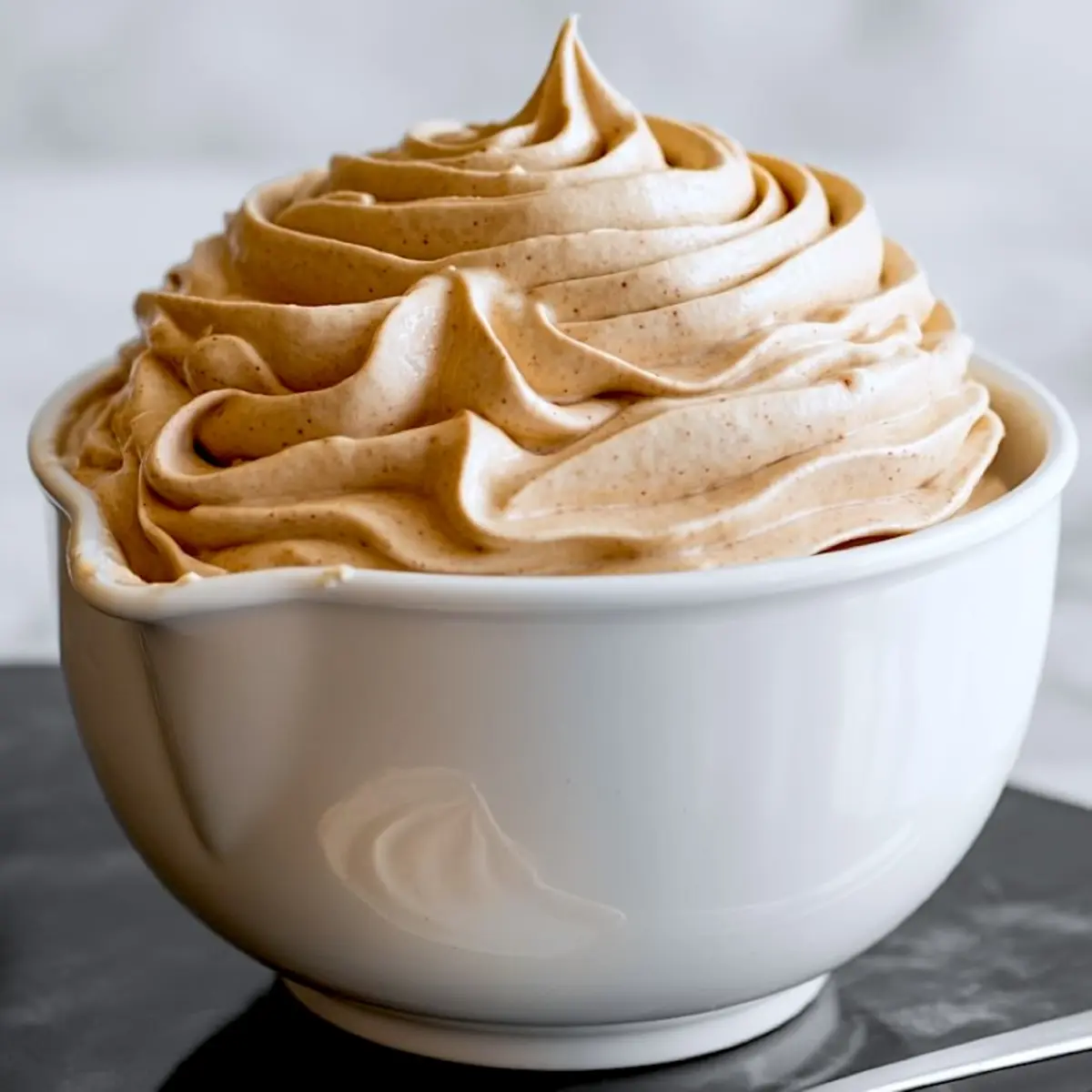 Creamy cinnamon buttercream frosting piped into elegant swirls in a white ceramic bowl, ready to top cupcakes or cakes.
