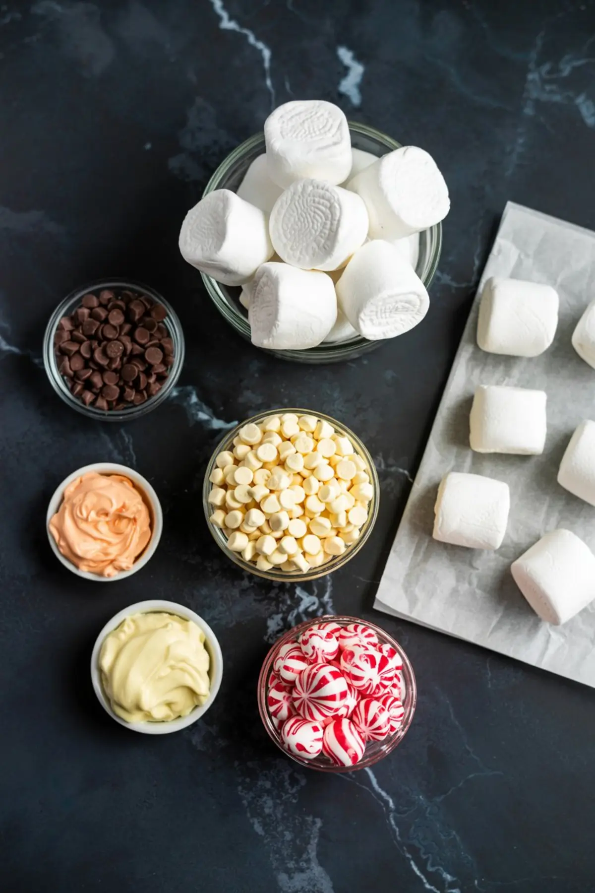 Flat lay of snowman cake pop ingredients including large marshmallows, white and dark chocolate chips, colored candy melts in orange and white, and peppermint candies, all arranged on a dark marble background with parchment paper.