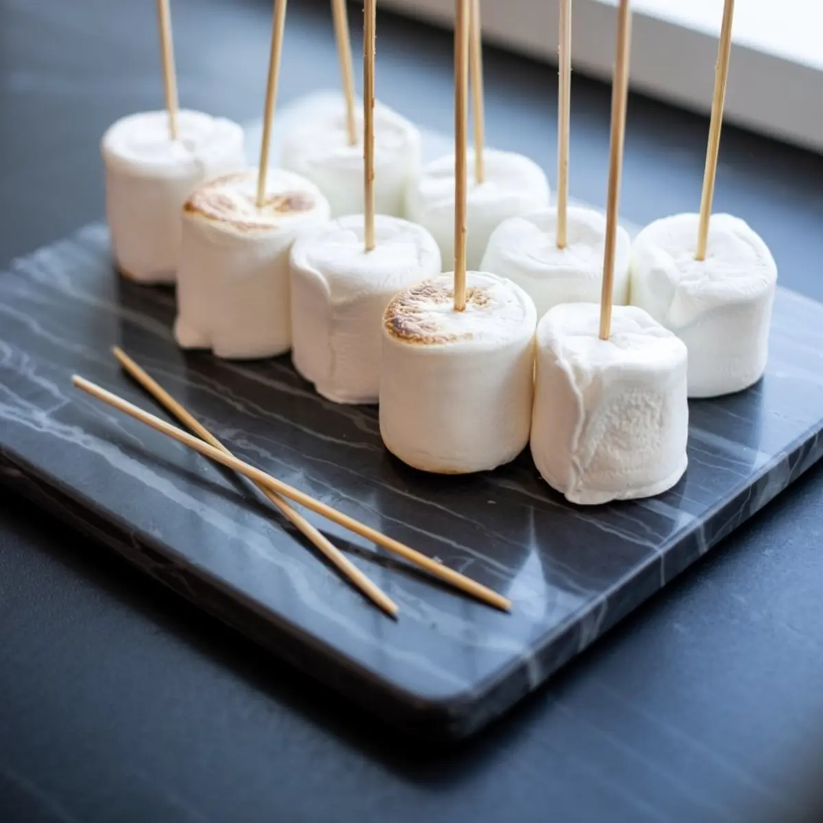 Preparation shot of skewered marshmallows arranged on a dark cutting board, with some marshmallows lightly toasted on top, ready to be dipped or decorated for making snowman cake pops.