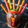 Close-up of a chocolate-dipped strawberry decorated as a turkey with candy eyes, orange beak, red wattle, and colorful pretzel stick feathers on a dark marble background.