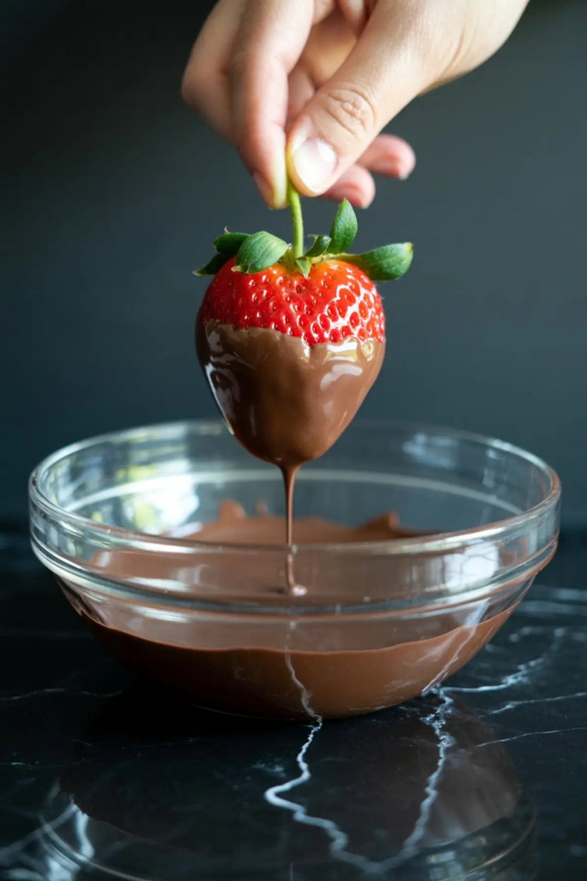 A hand dipping a ripe strawberry into a glass bowl of melted chocolate, with chocolate dripping from the fruit.