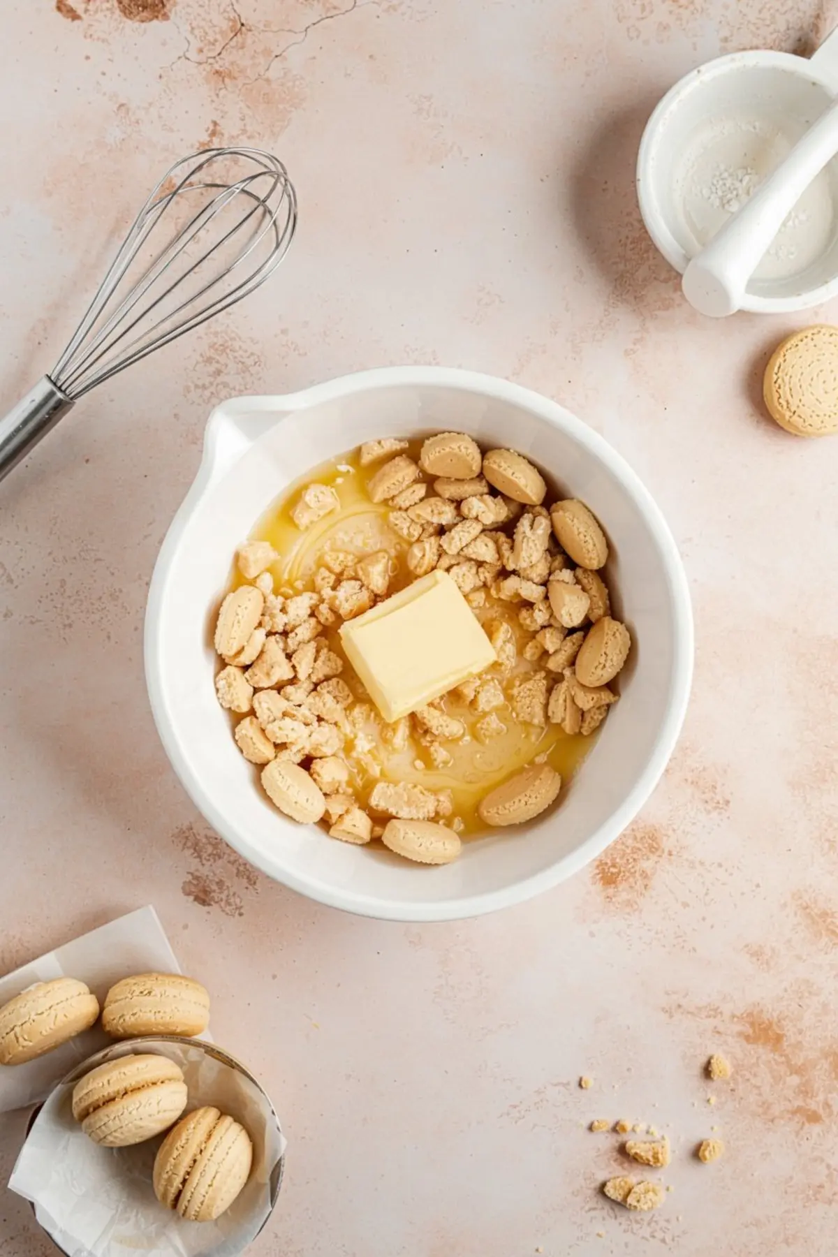 Bowl of crushed vanilla sandwich cookies mixed with melted butter for cheesecake crust preparation, surrounded by whole cookies and baking tools on a light pink countertop.