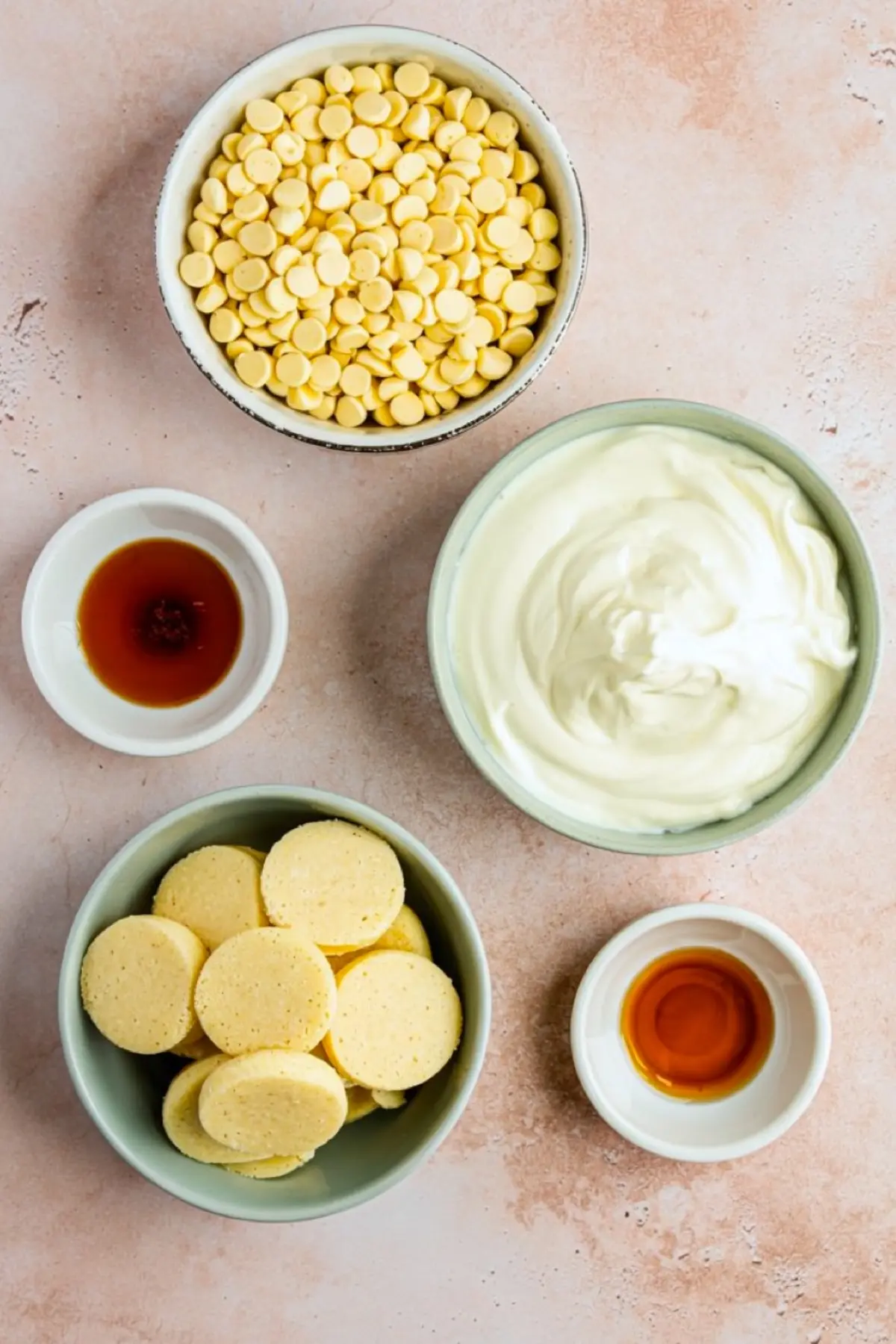 Flat lay of sugar cookie fudge ingredients including white chocolate chips, whipped topping, vanilla extract, sugar cookies, and sweetened condensed milk, displayed in small bowls on a light surface.
