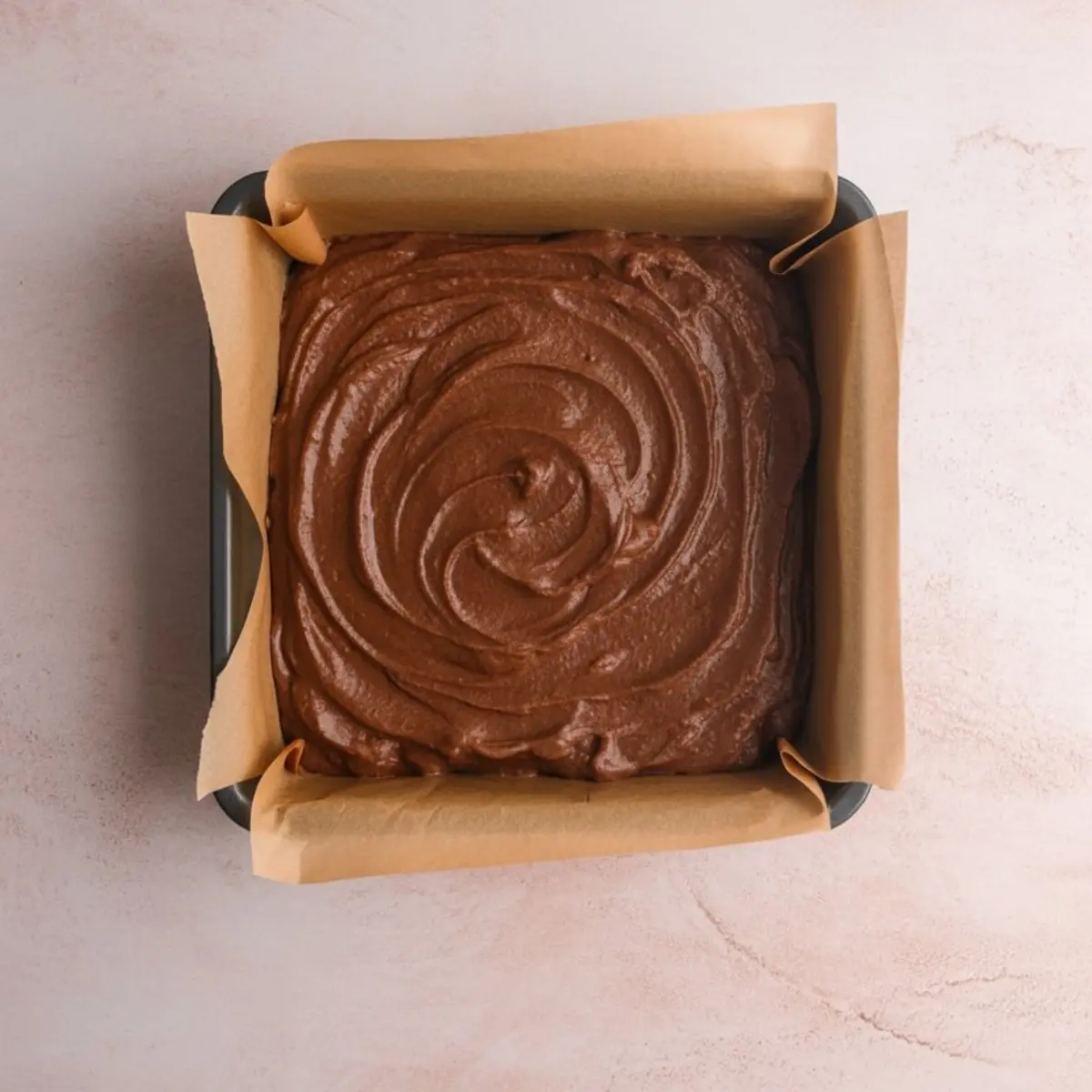 Unbaked sweet potato brownie batter spread evenly in a square baking tin lined with parchment paper. The smooth, swirled top surface reflects the creamy, rich chocolate blend before baking.