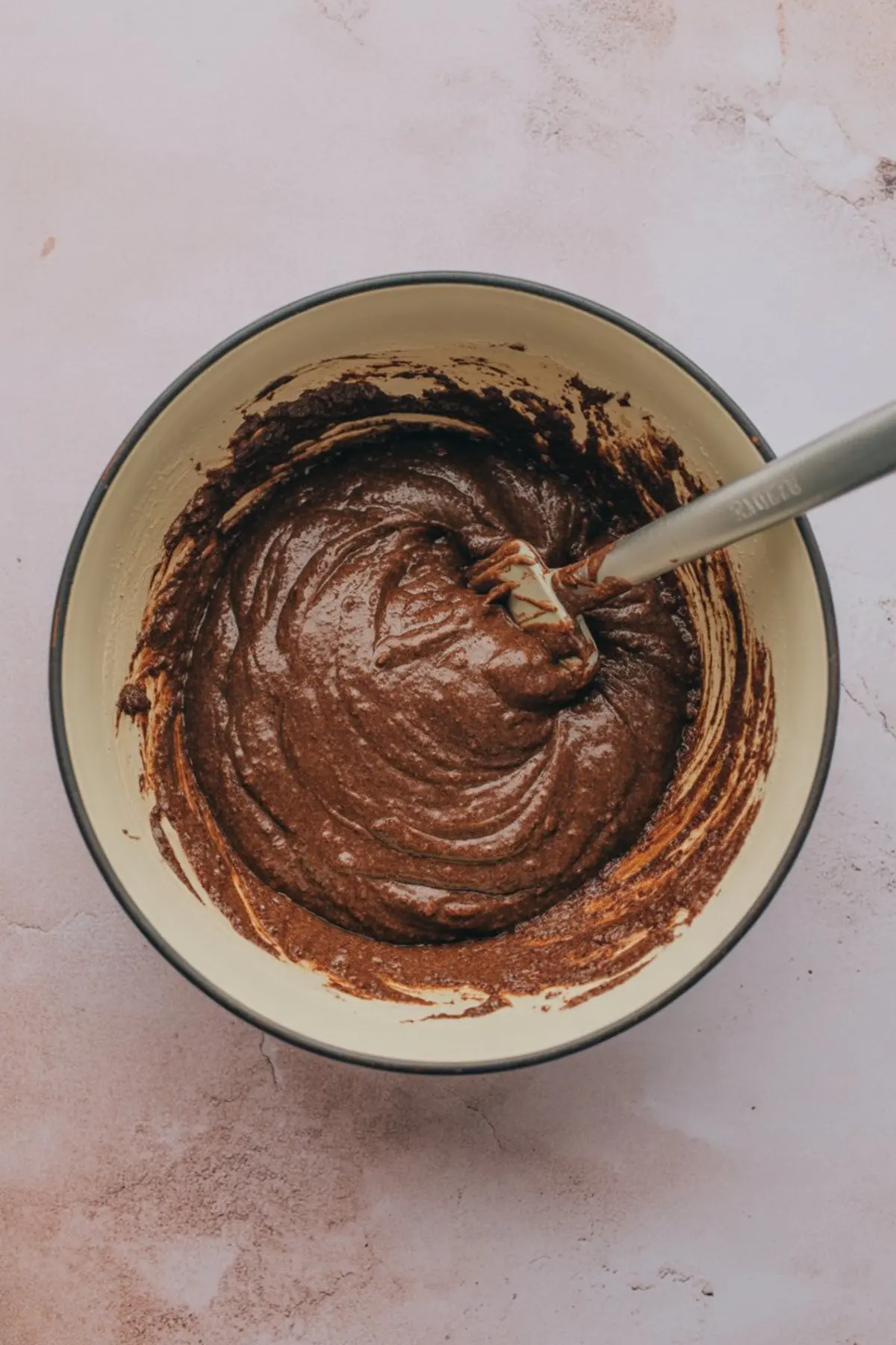 Overhead view of sweet potato brownie batter in a mixing bowl with a spatula resting inside. The thick, chocolatey mixture clings to the sides, capturing the rich consistency and freshly mixed texture.