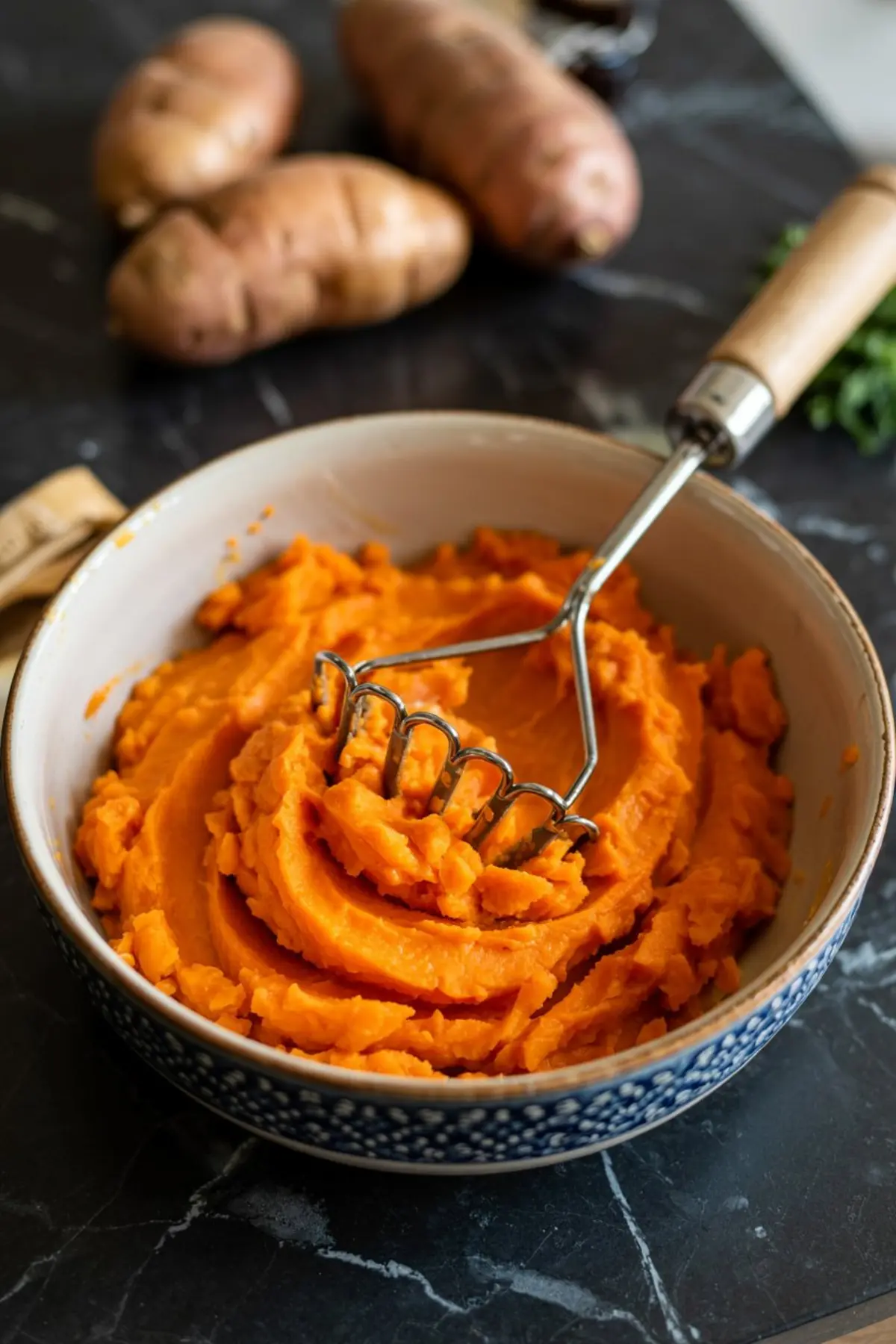 Bowl of mashed sweet potatoes with a metal masher, showing vibrant orange texture on a dark countertop, ideal for sweet potato recipes and Thanksgiving dishes.