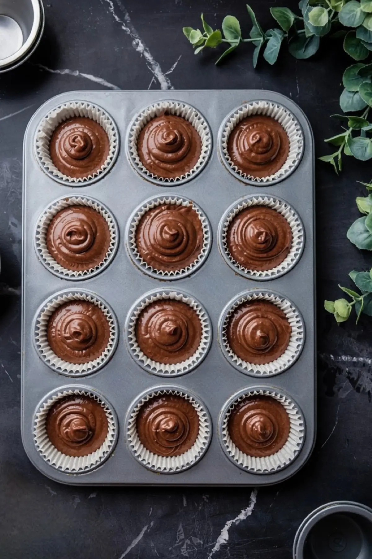 Twelve cupcake liners filled with chocolate batter in a metal muffin tin, ready to be baked, placed on a dark marble surface.
