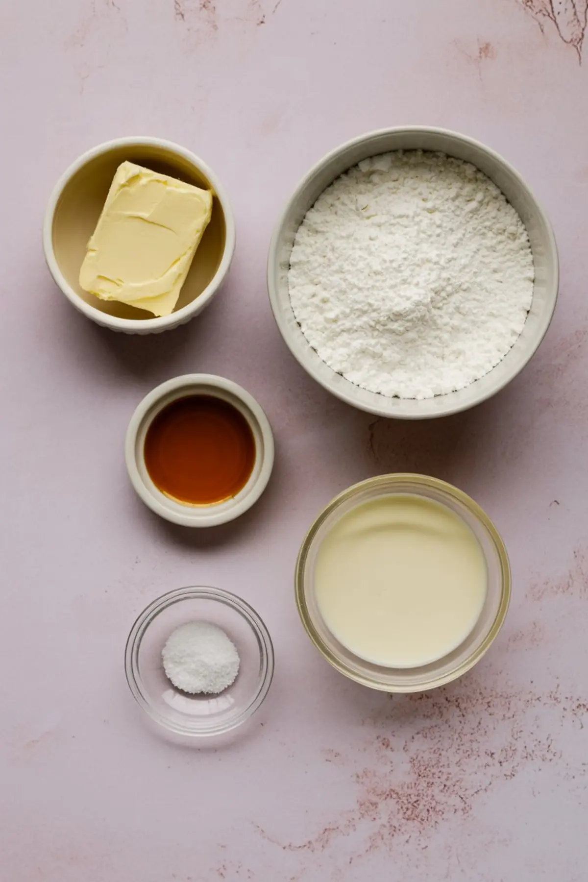 Flat lay of buttercream frosting ingredients including unsalted butter, powdered sugar, heavy cream, vanilla extract, and salt arranged in ceramic and glass bowls on a light pink surface.