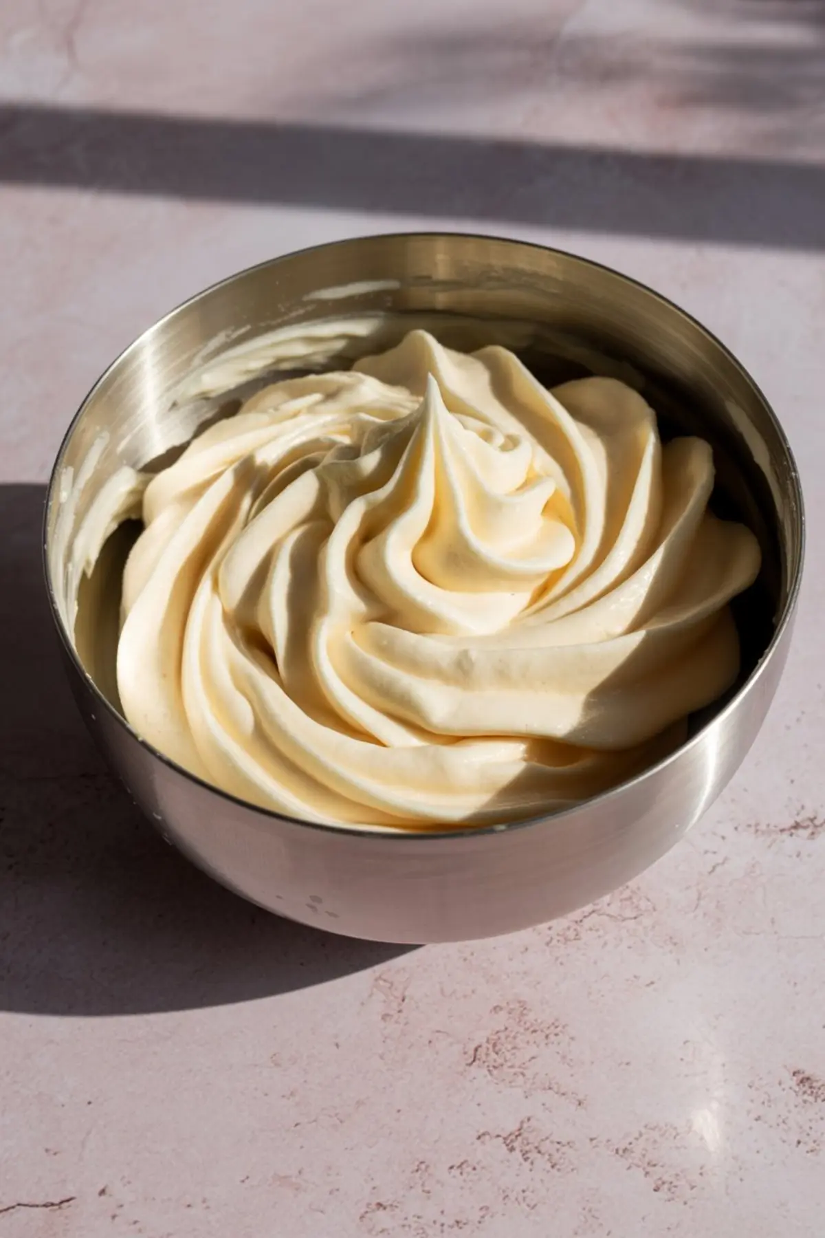 Fluffy vanilla buttercream frosting with soft peaks swirled in a stainless steel mixing bowl, sitting on a pink countertop under natural light.