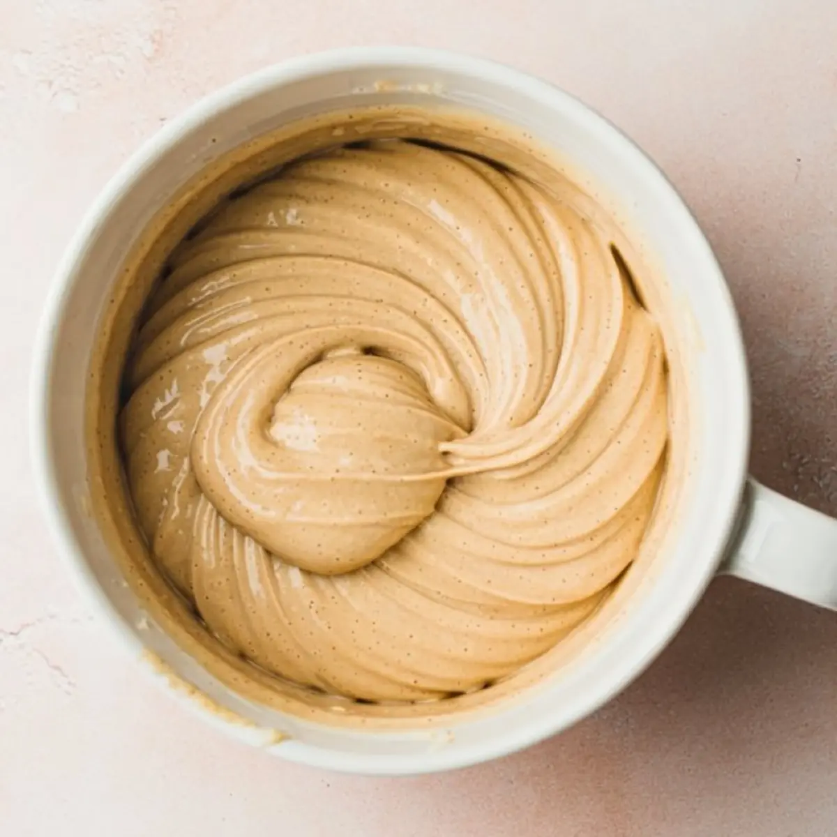Creamy vanilla cake batter swirled in a white ceramic mug, showing a smooth, thick texture with a rich golden color on a soft pink background.