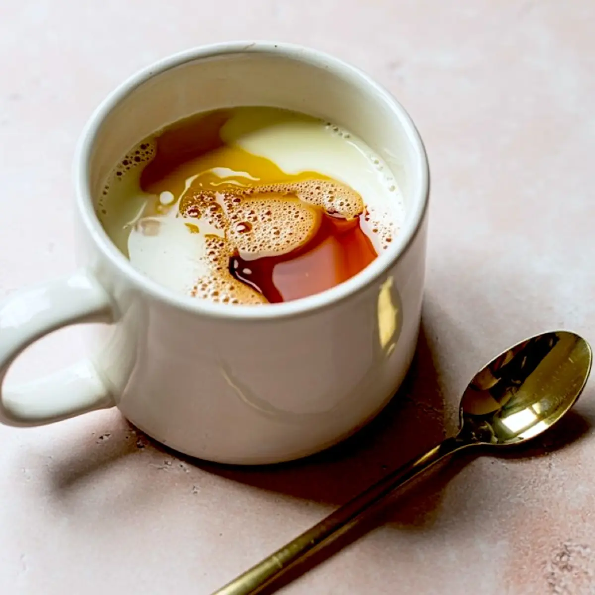 Overhead view of a white mug filled with milk, vanilla extract, and oil before mixing, placed beside a gold spoon on a pastel pink background.