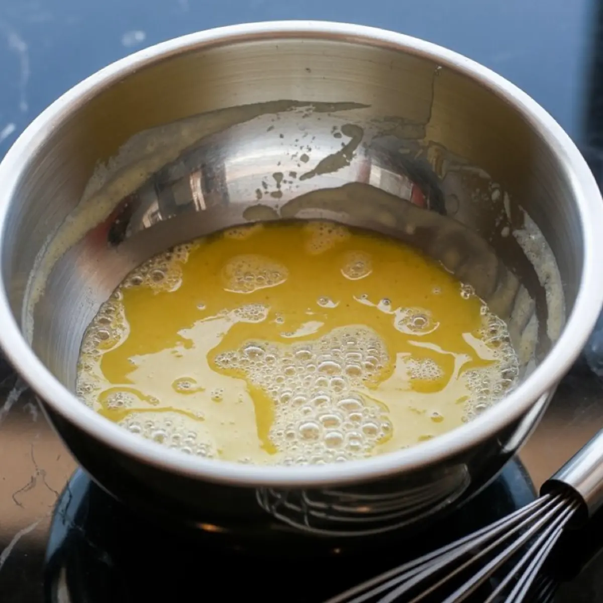 Melted butter and sugar mixture in a stainless steel mixing bowl with a whisk resting beside it on a black countertop.
