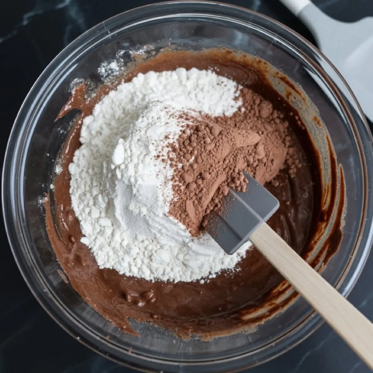 Flour and cocoa powder being folded into a chocolate batter using a rubber spatula in a glass mixing bowl.
