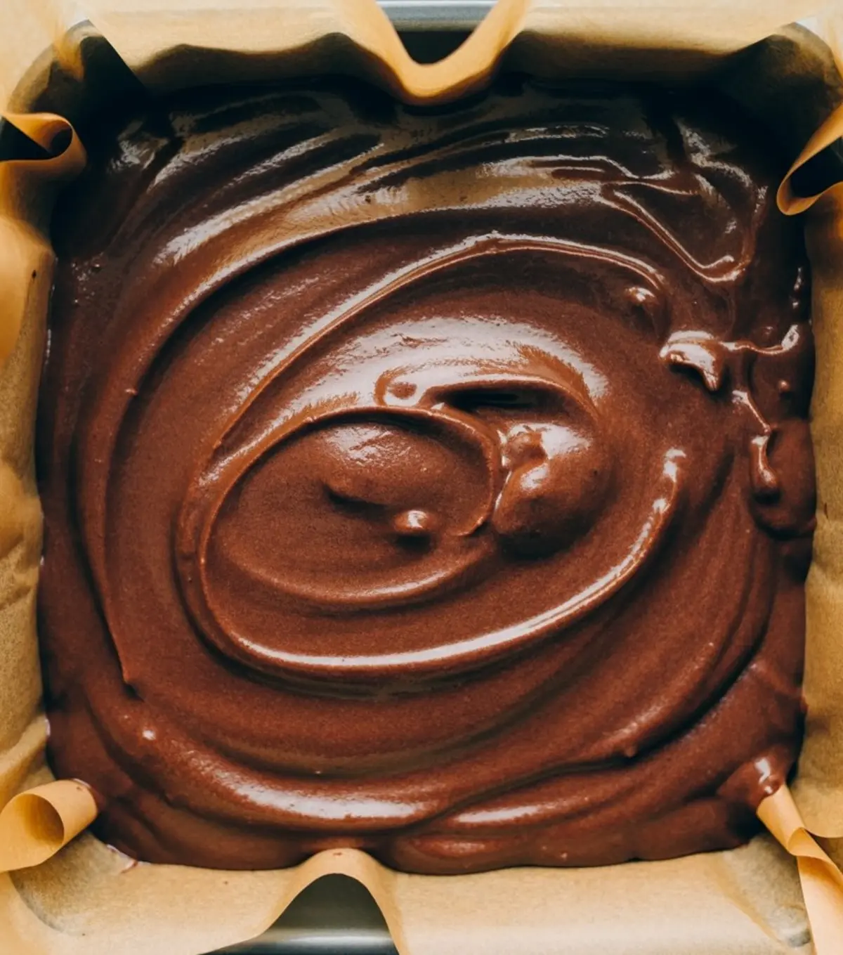 Raw brownie batter evenly spread in a parchment-lined square baking pan, ready to be baked.
