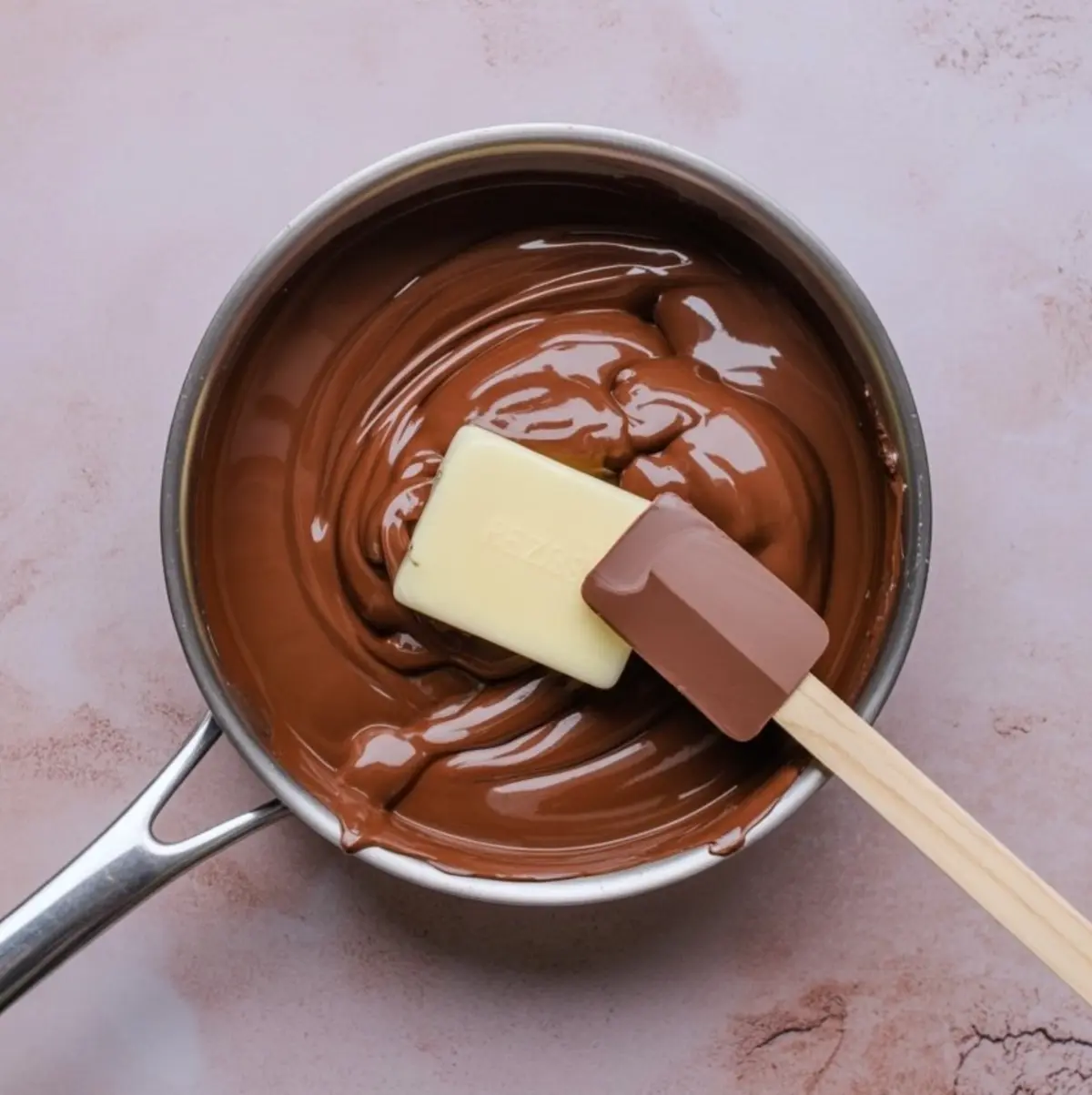 Melted chocolate in a saucepan being stirred with a white spatula and a brown spatula, resting on a pink surface.
