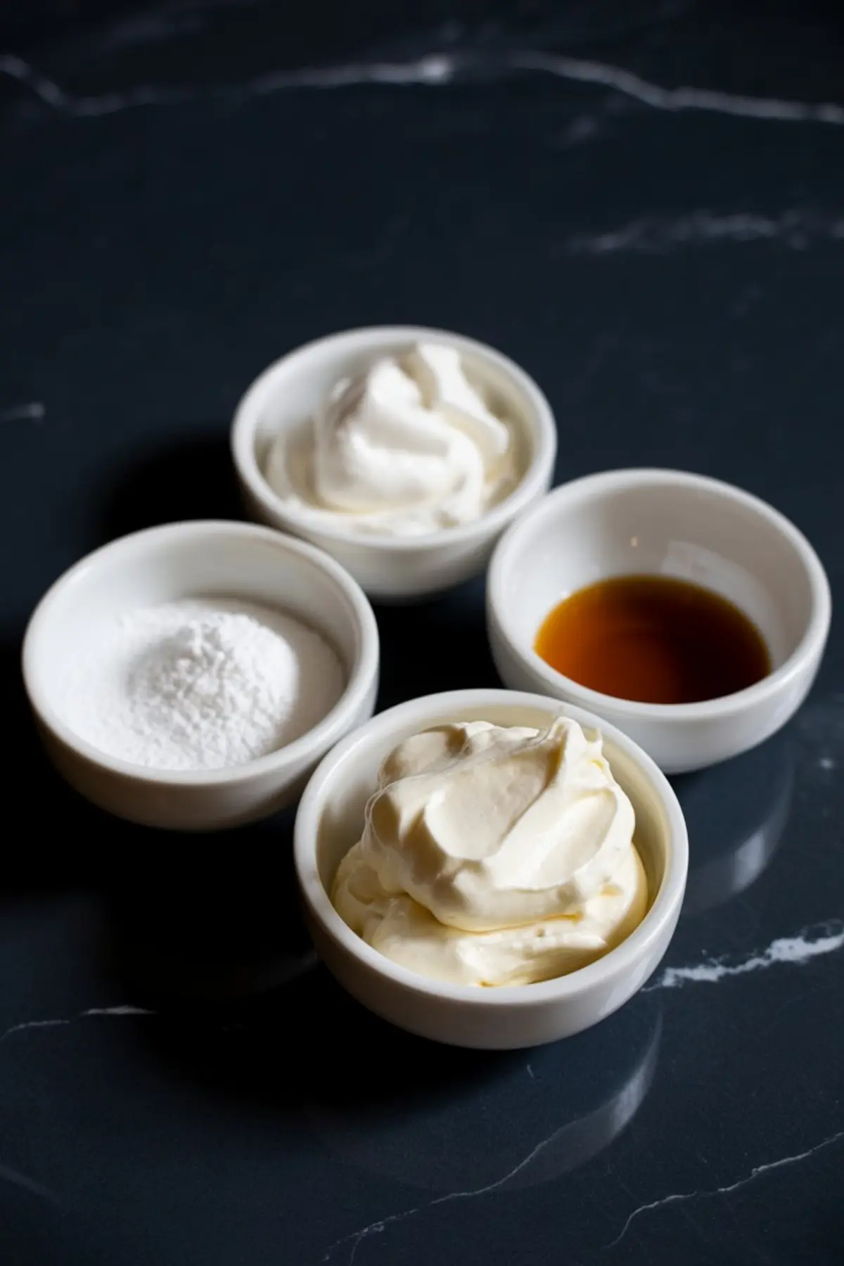 Four small white bowls hold whipped cream, powdered sugar, vanilla extract, and mascarpone cheese, displayed on a dark marble surface.