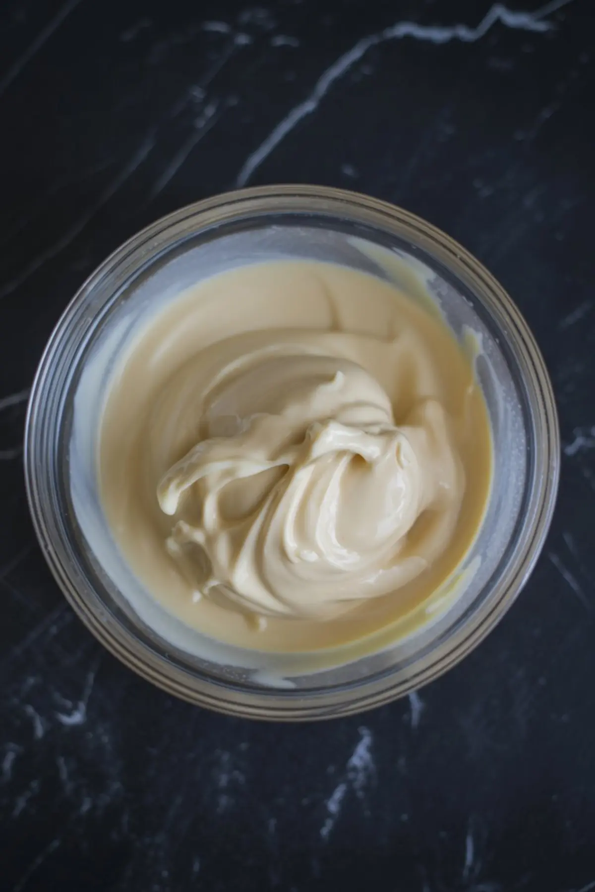 Creamy white chocolate frosting in a glass bowl on a dark marble surface, showing a smooth and rich whipped texture.