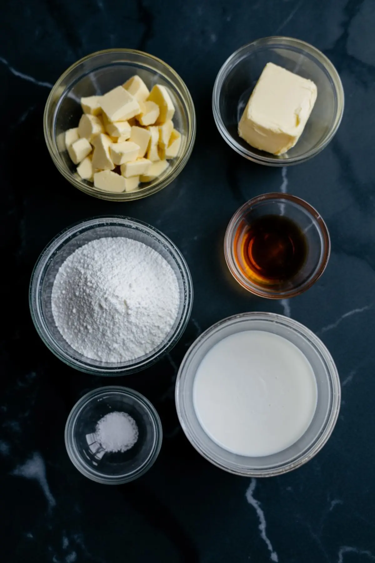Overhead view of measured ingredients for white chocolate frosting including white chocolate chunks, butter, powdered sugar, vanilla extract, cream, and salt in glass bowls.
