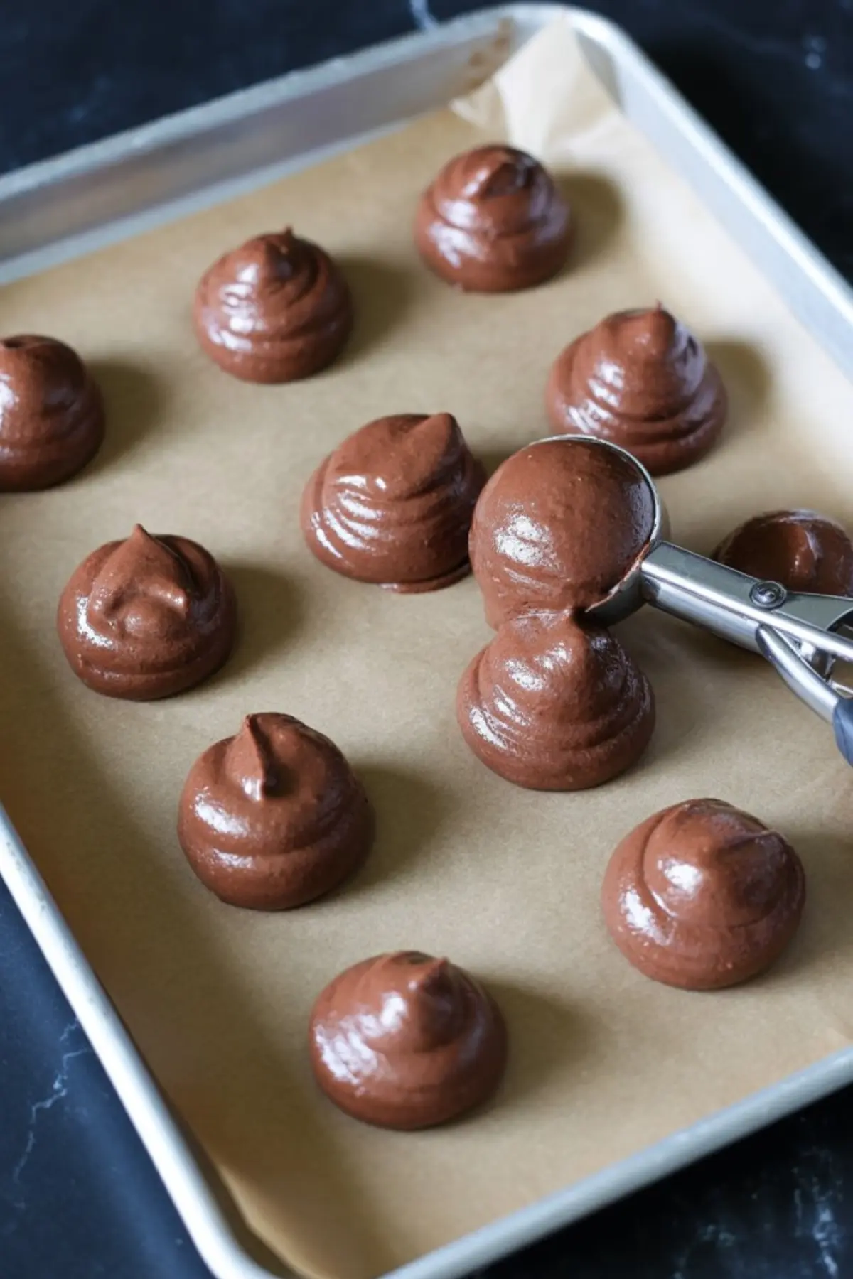 Chocolate whoopie pie batter scooped into round portions on a parchment-lined baking sheet, with a cookie scoop in action.