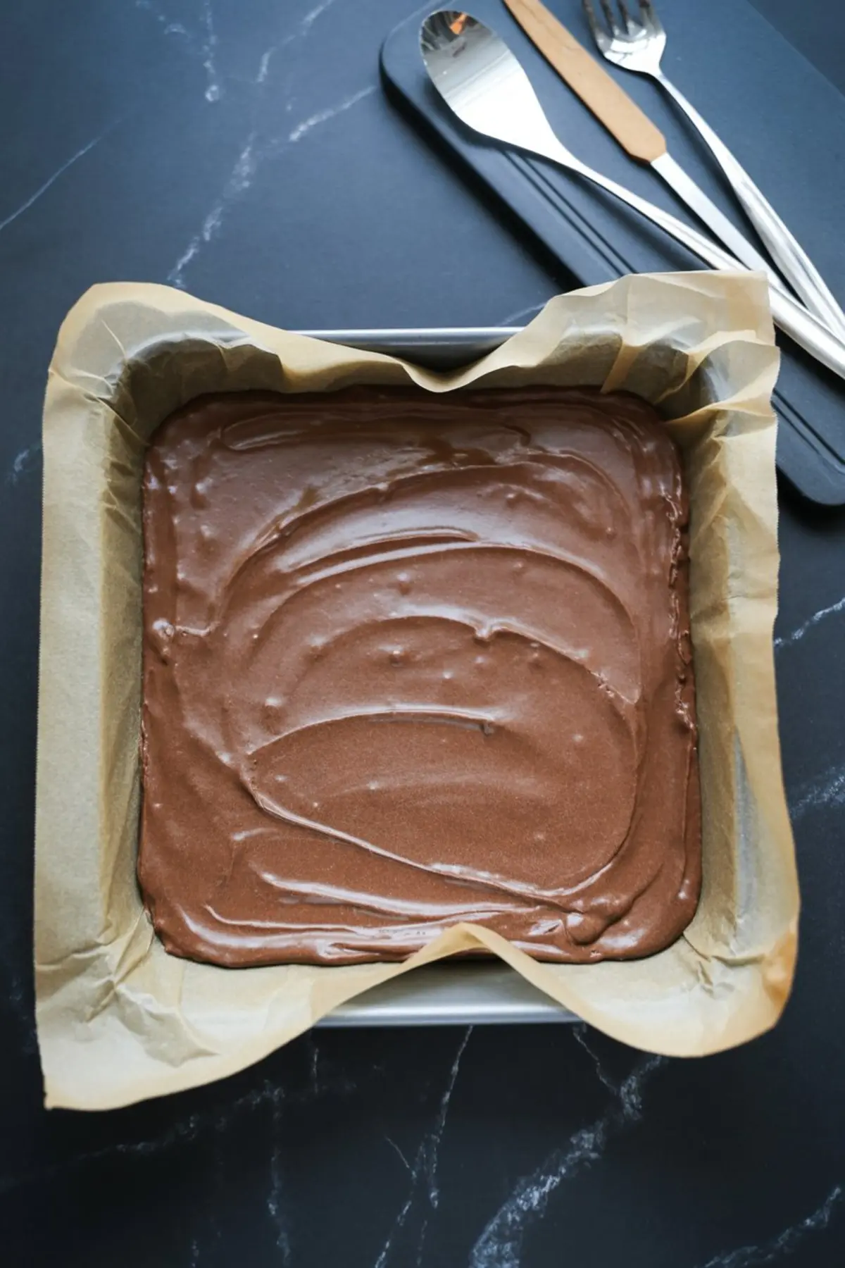 Chocolate brownie batter spread evenly in a parchment-lined square baking pan, prepared for the base layer of dessert bars.