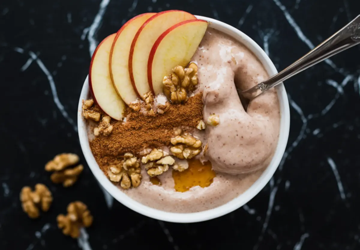 Apple cinnamon smoothie bowl topped with thin apple slices, crushed walnuts, ground cinnamon, and a drizzle of honey, served in a white bowl with a spoon.