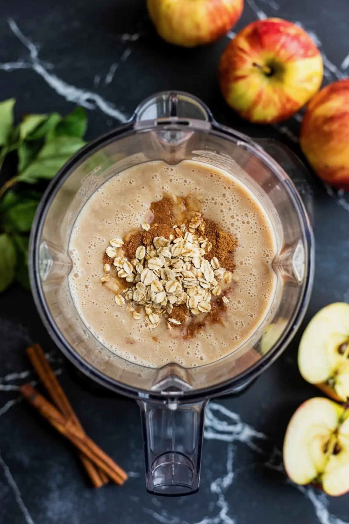 Top view of a blender filled with apple cinnamon smoothie ingredients, including rolled oats, ground cinnamon, and fresh apple chunks on a black marble countertop.