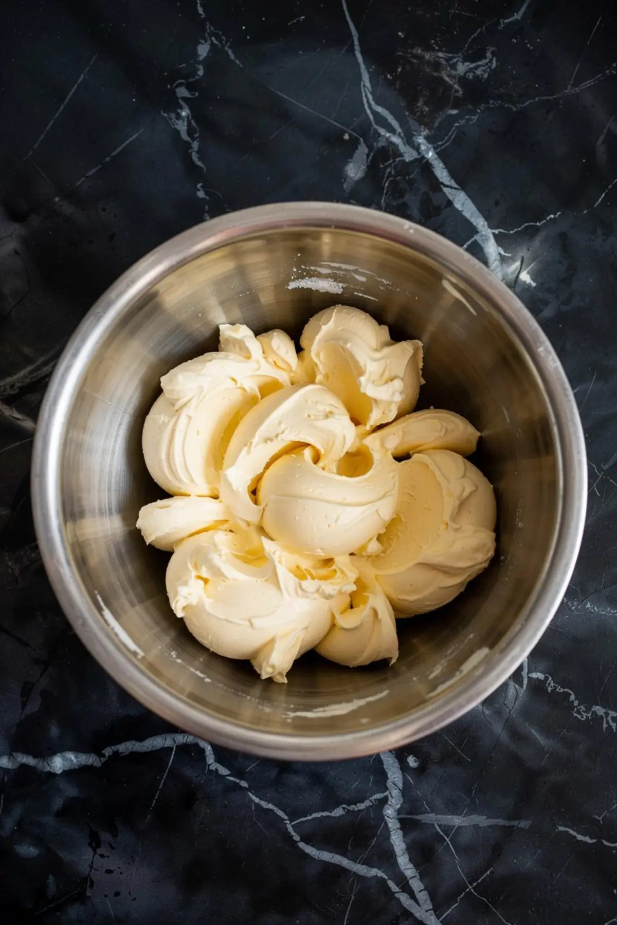Whipped butter in a metal bowl, featuring smooth swirls of pale yellow cream, as the starting base for homemade chocolate frosting.