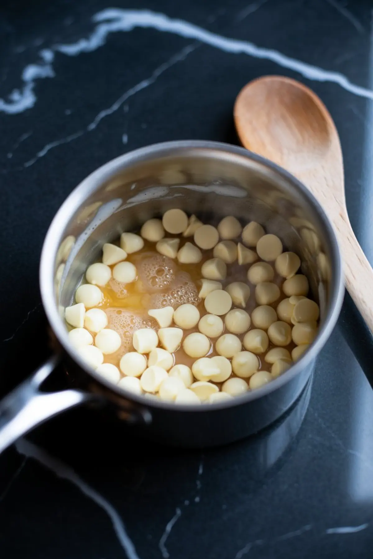 Close-up of a saucepan with melting white chocolate chips and butter on a black marble surface, alongside a wooden spoon.
