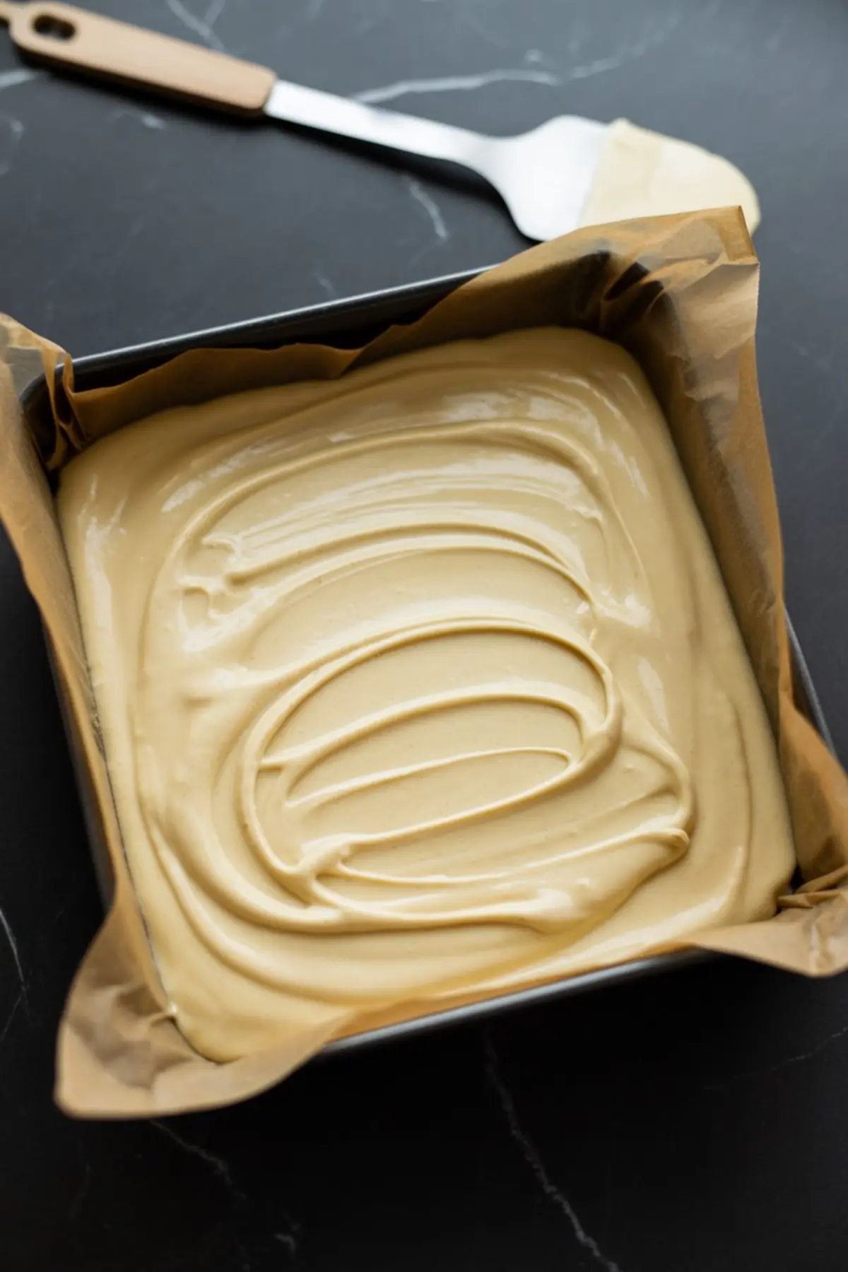 Square baking pan lined with parchment paper filled with smooth, swirled white fudge mixture, ready to set.
