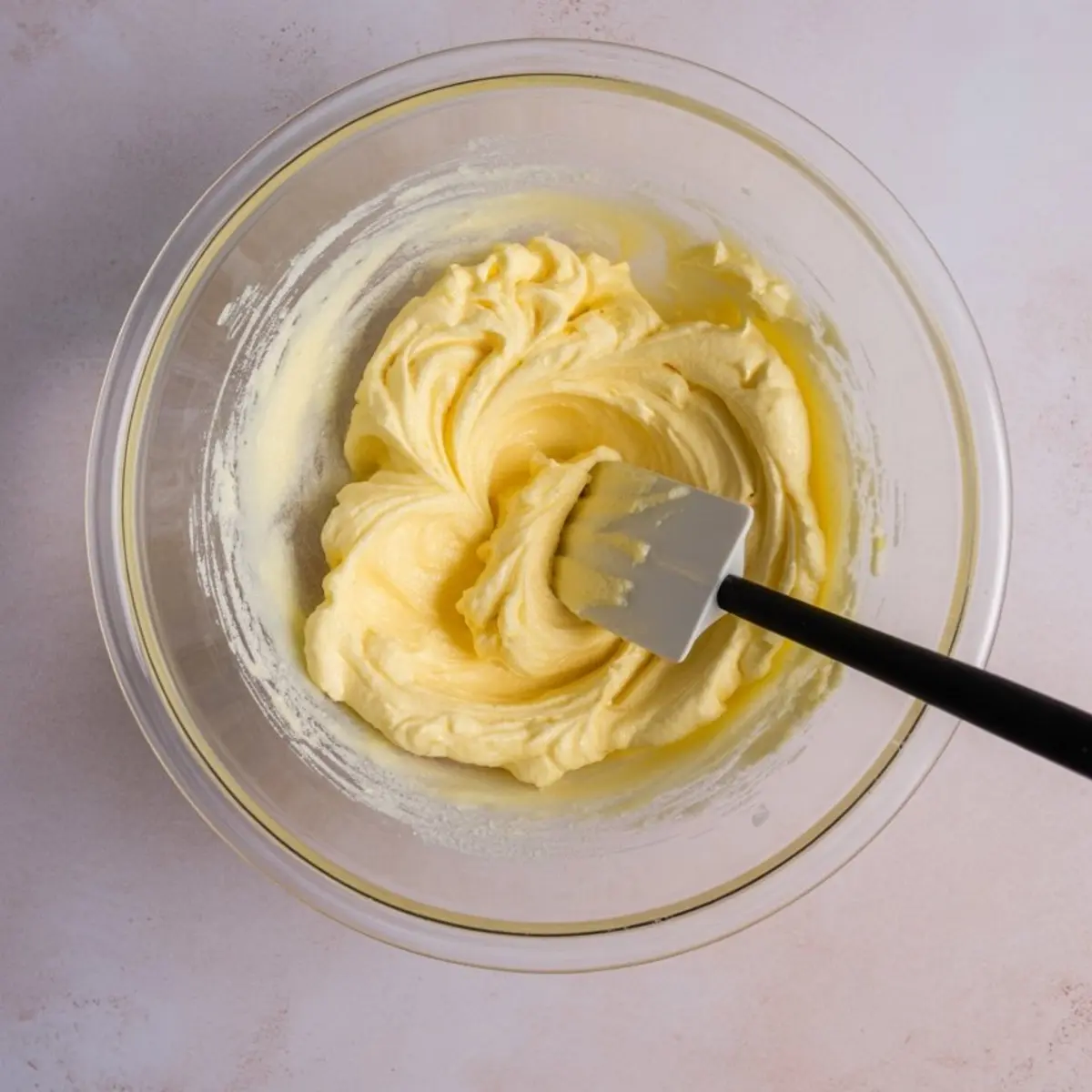 Glass mixing bowl filled with creamy yellow cake batter being stirred with a black-handled spatula on a pink countertop.