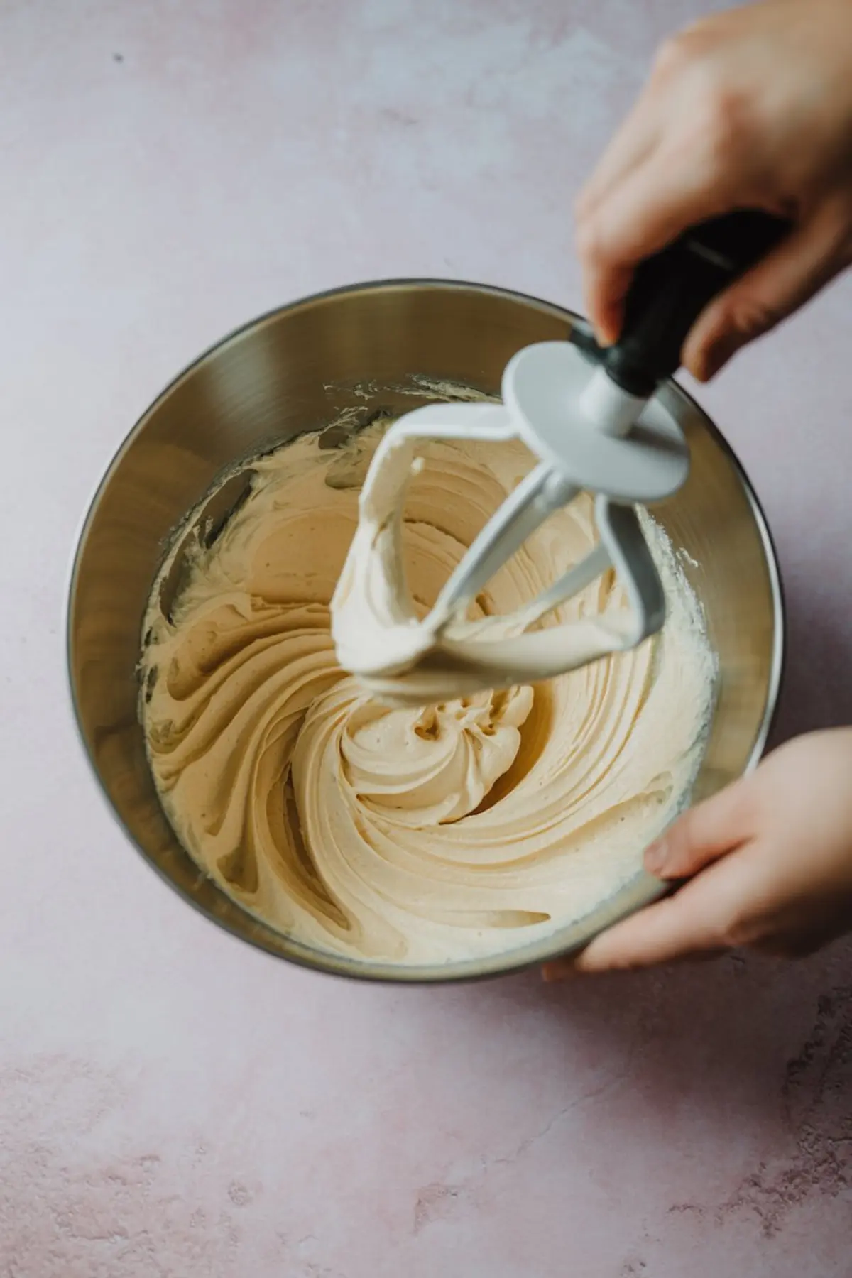 Overhead view of thick vanilla cake batter in a metal mixing bowl with a stand mixer paddle attachment resting on the side, held by two hands.