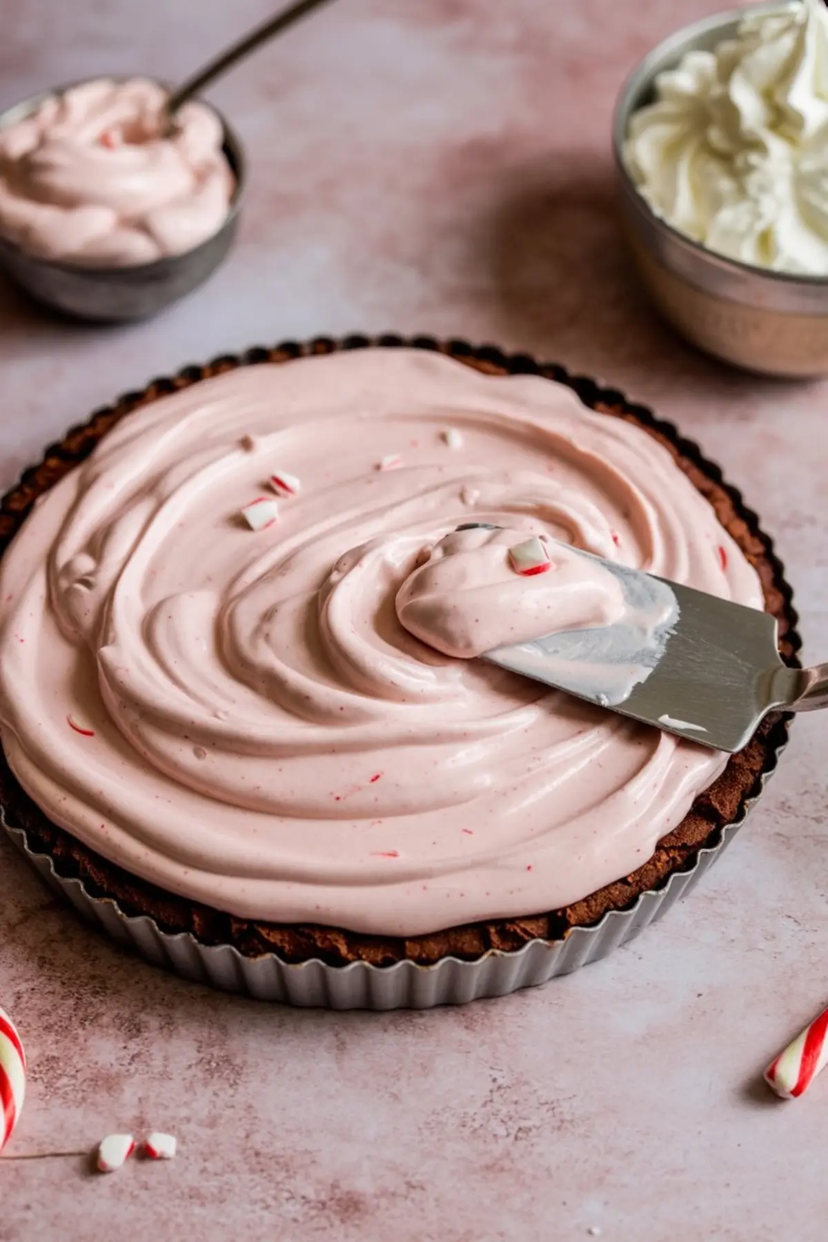 Candy cane pie in a fluted chocolate crust, pink peppermint cream swirled with an offset spatula, tiny crushed candy pieces resting on the surface.