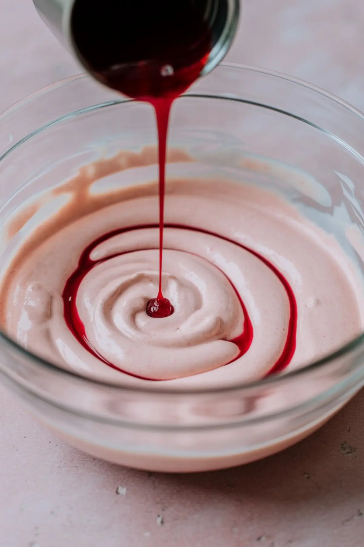 Thin stream of deep red peppermint syrup pouring into creamy pink mixture, forming a spiral in a clear mixing bowl.