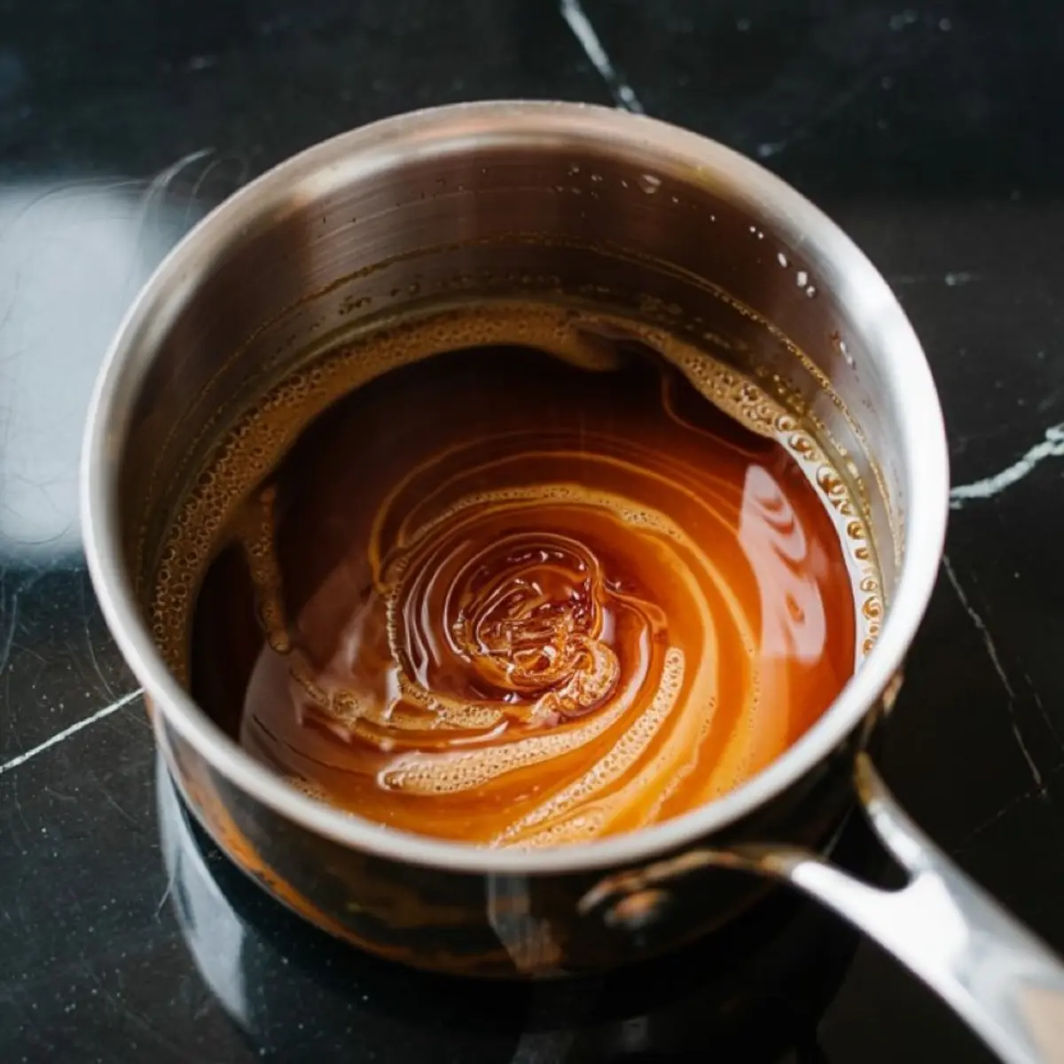 Golden caramel sauce swirled in a stainless steel saucepan, showcasing its glossy texture and rich amber color on a black marble surface.
