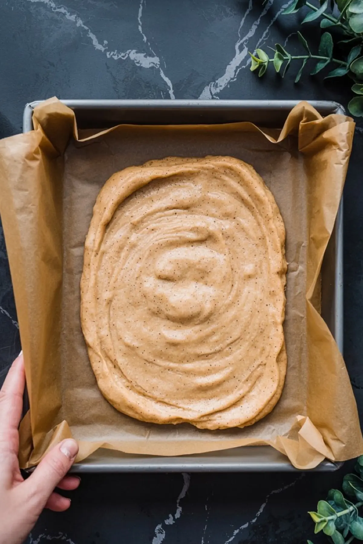 Baking tray lined with parchment paper holding an unbaked snickerdoodle cheesecake swirl layer, showing a smooth cinnamon-speckled batter spread in an oval shape on a dark kitchen countertop.