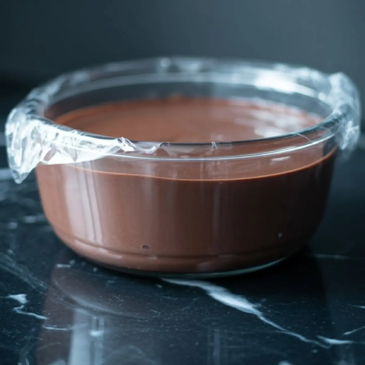 Glass bowl covered with plastic wrap holding silky chocolate custard set on a black marble counter.
