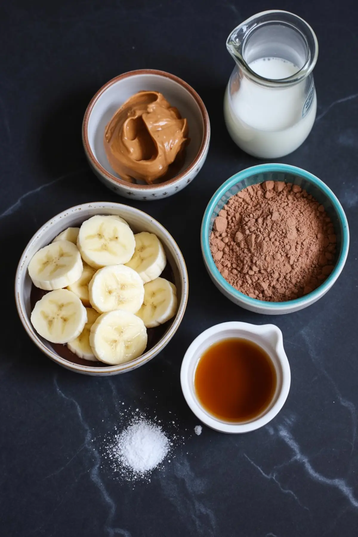 Flat lay of smoothie ingredients including sliced bananas, peanut butter, cocoa powder, vanilla extract, milk, and a pinch of salt on a black marble background.