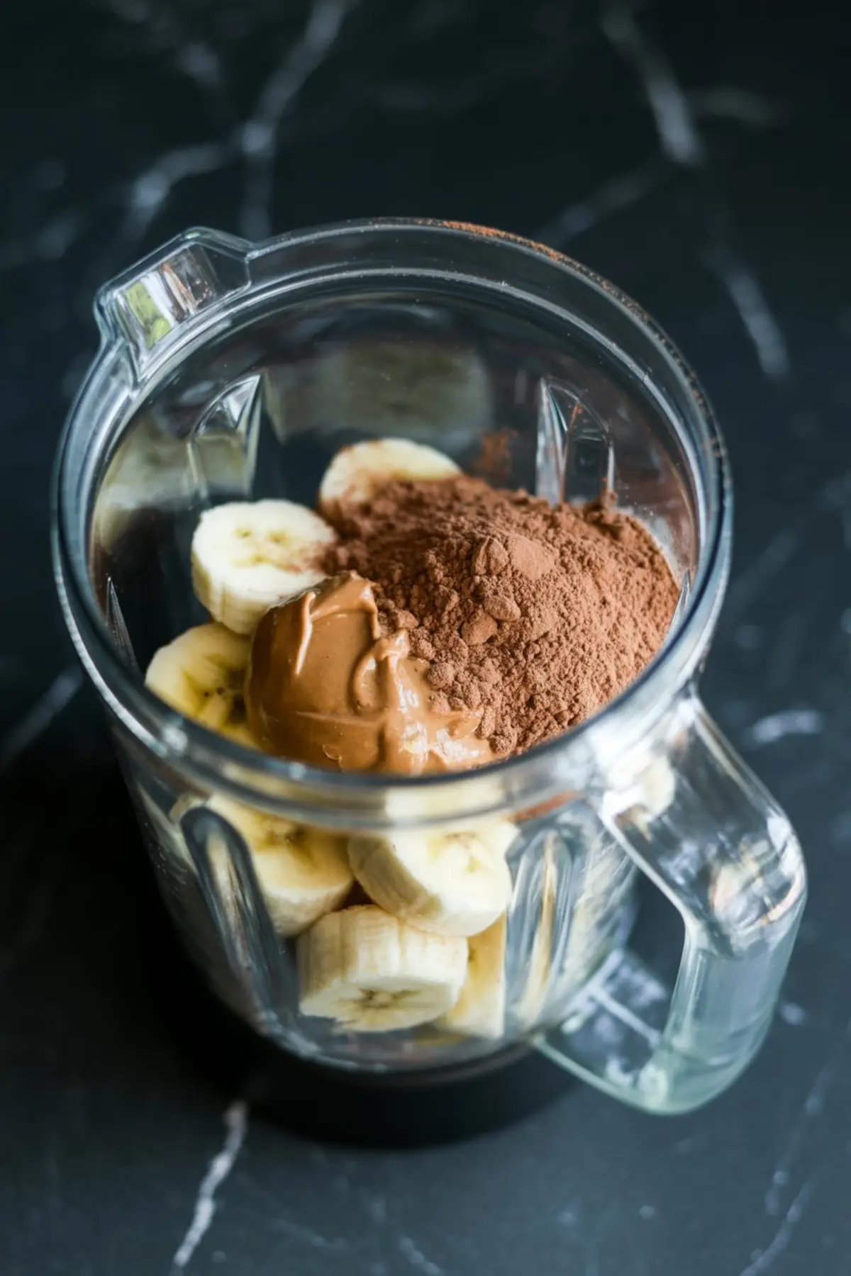 Blender filled with sliced bananas, peanut butter, and cocoa powder before blending, captured from a top-down angle.