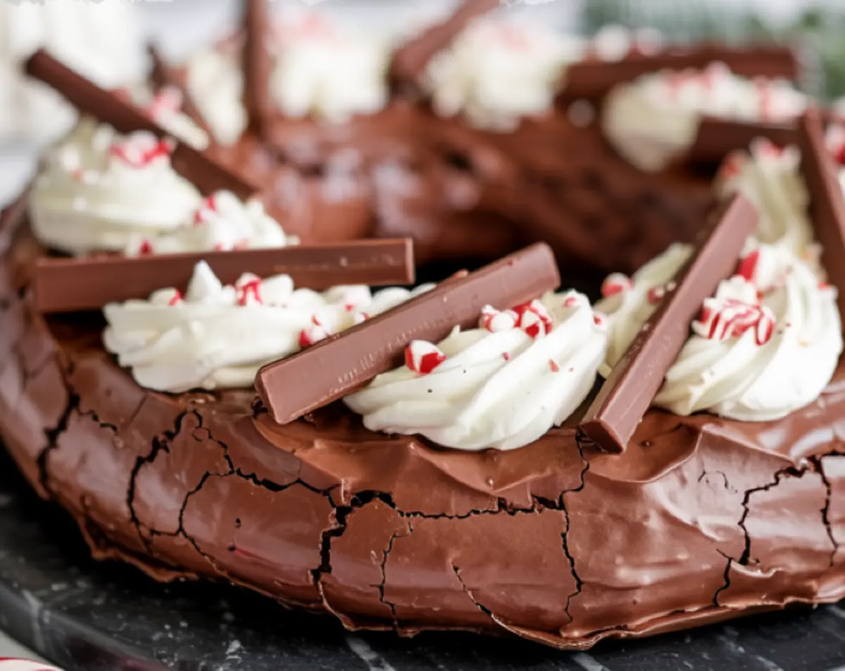 Close-up side view of chocolate peppermint pavlova wreath showing cracked meringue texture, whipped cream swirls, crushed peppermint, and chocolate bars.
