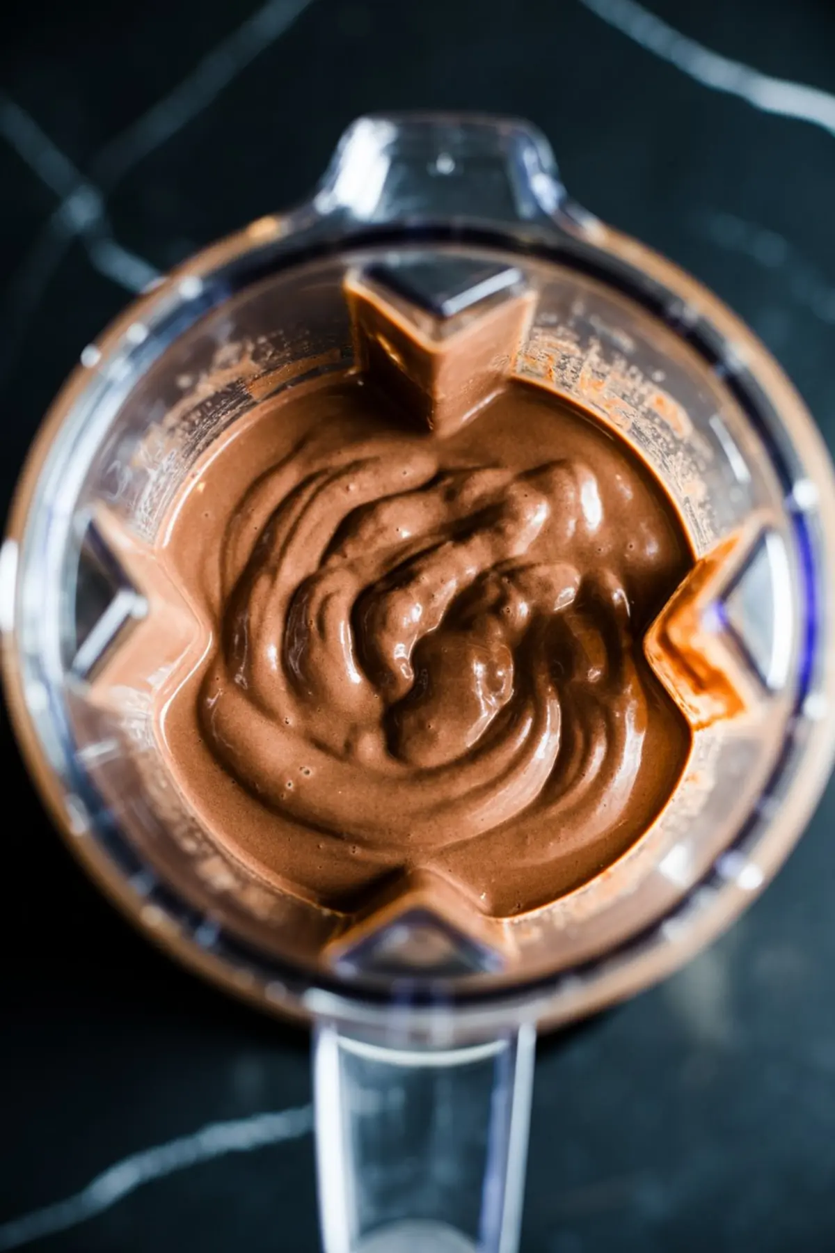 Top view of thick chocolate smoothie blended to a smooth consistency inside a clear blender container, sitting on a dark marble surface.