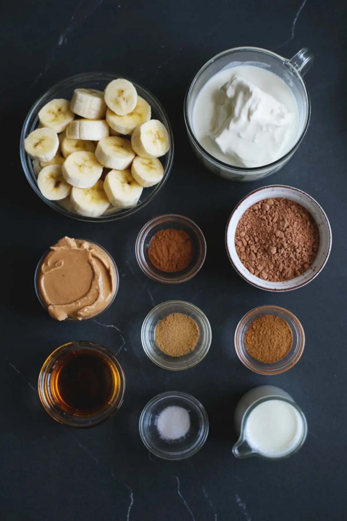 Flat lay of smoothie ingredients including sliced bananas, Greek yogurt, peanut butter, cocoa powder, cinnamon, vanilla extract, cardamom, salt, and milk on a black stone background.