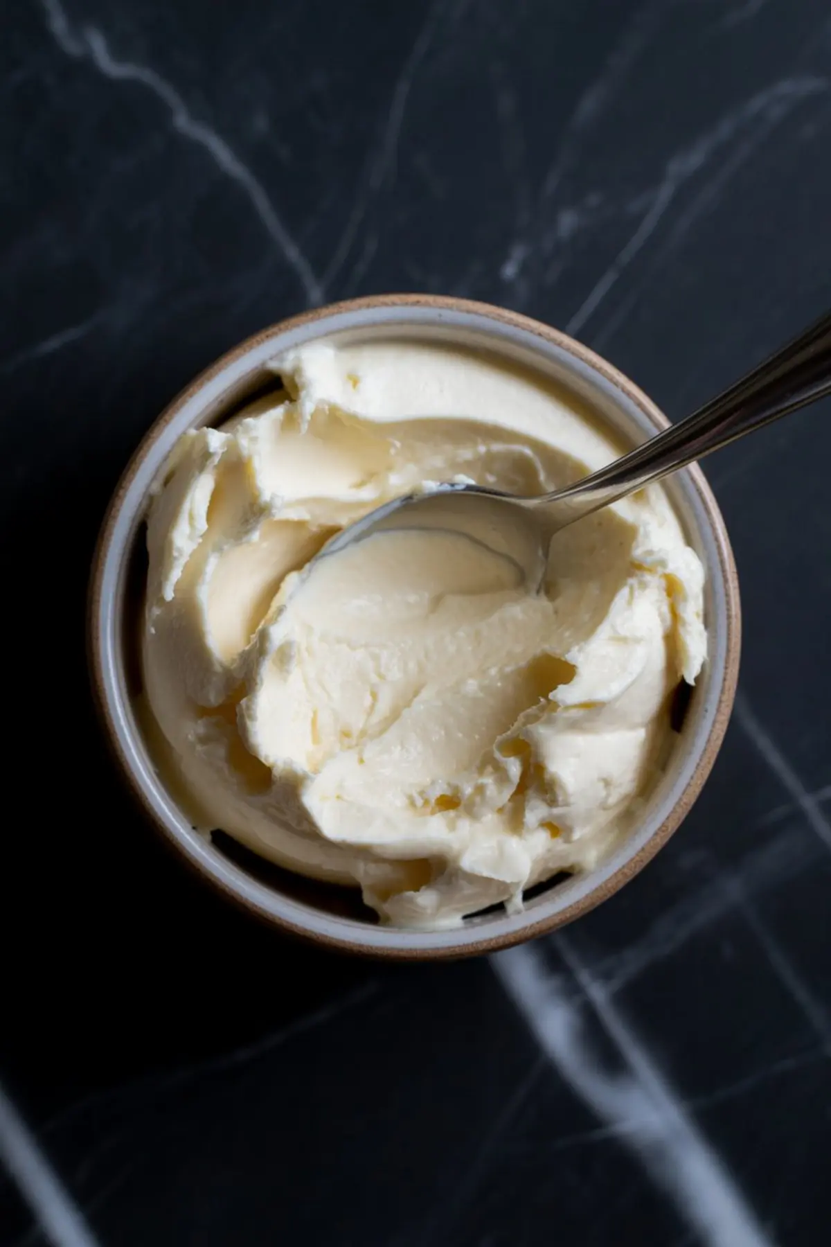 Creamy vanilla frosting in a ceramic bowl with a spoon, showcasing its thick and smooth texture on a dark marble surface.
