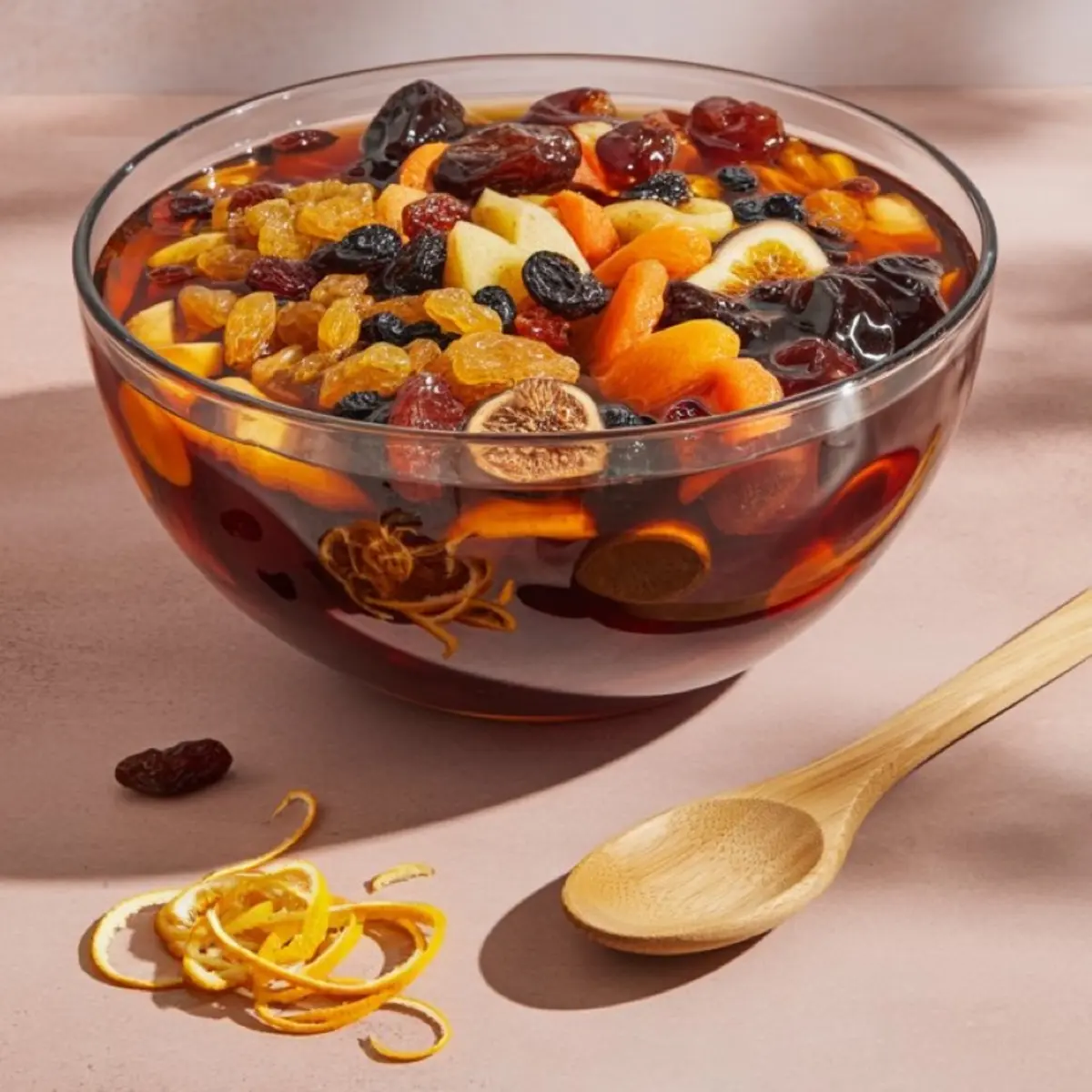 Glass bowl filled with dried apricots, figs, raisins, and citrus peel soaking in dark liquor, prepared for making traditional fruit cake.
